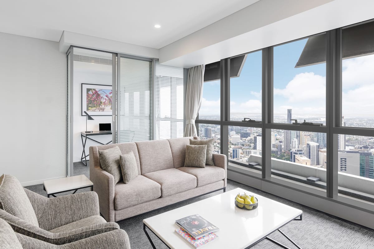 A modern living room with a large window offering a city view, a beige sofa, a small table with a magazine, and a coffee table with fruit.