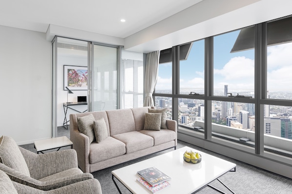 Una sala de estar moderna con un ventanal que ofrece vistas a la ciudad, un sofá color beige, una mesita con una revista y una mesa de centro con fruta.