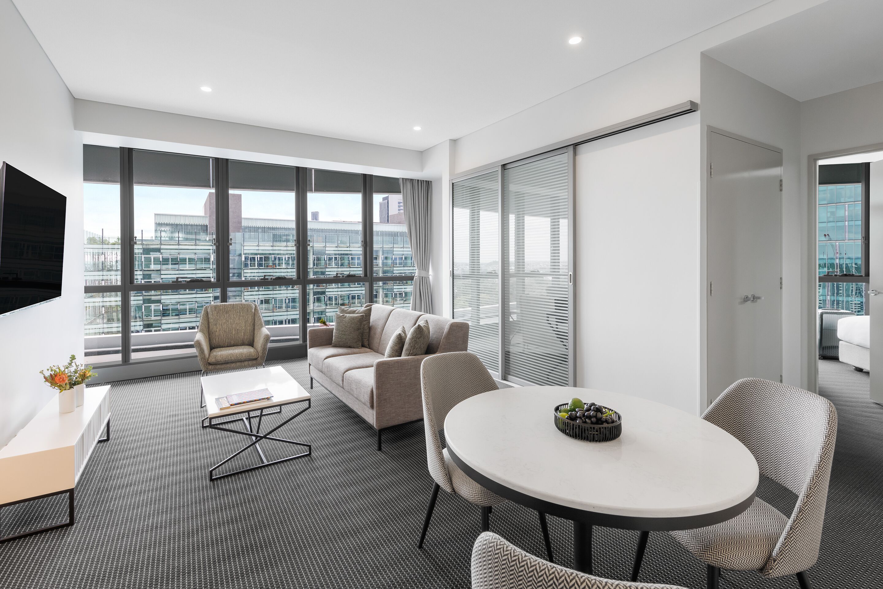 A modern hotel room with a large window, a sofa, a dining table, and a TV.