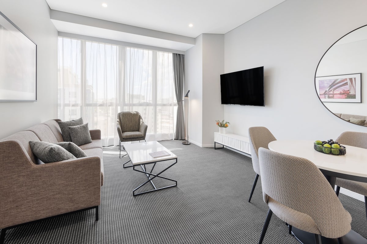 A modern hotel room with a sofa, armchair, dining table, and a flat-screen TV mounted on the wall.