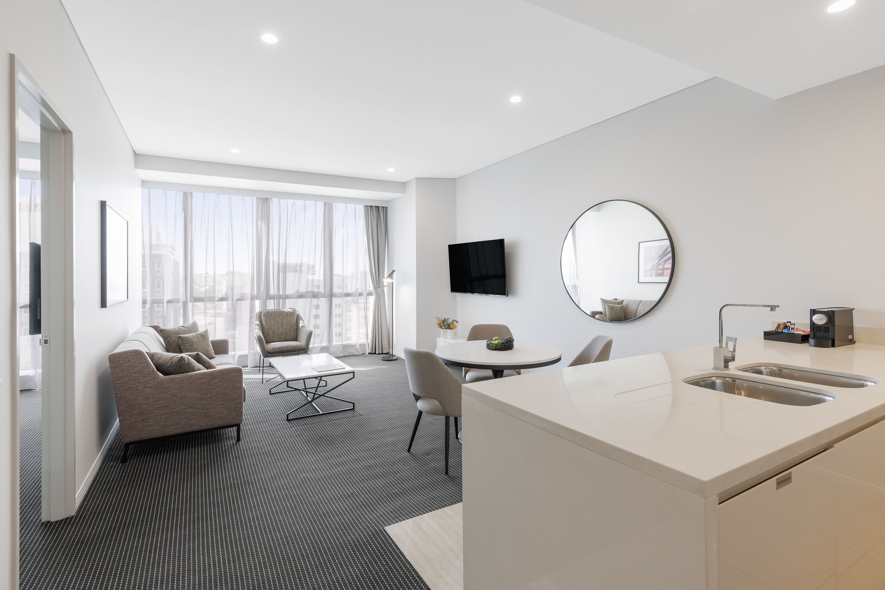 A modern hotel room with a sofa, dining table, chairs, a kitchen area with a sink, a flat-screen TV, and large windows offering a city view.