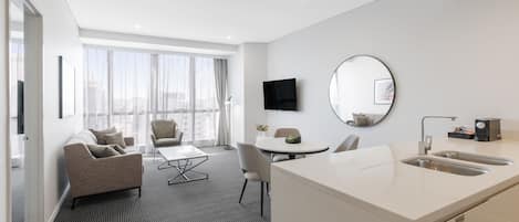 Una habitación de hotel moderna con sofá, mesa de comedor, sillas, cocina con lavamanos, televisor de pantalla plana y ventanas amplias con vistas a la ciudad.