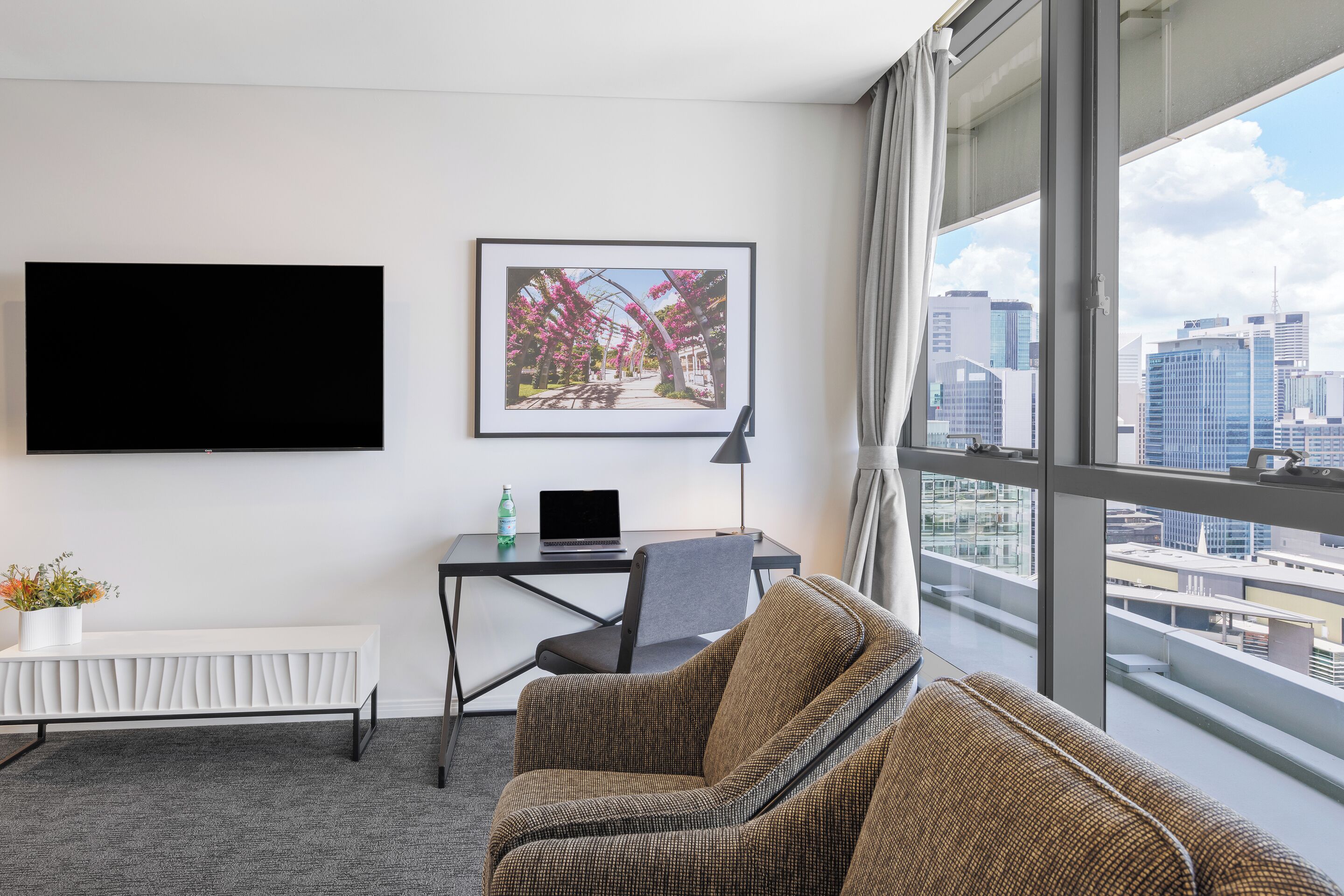 A modern hotel room with a large window, a flat-screen TV, a desk with a laptop, a wicker sofa, and a framed picture on the wall.