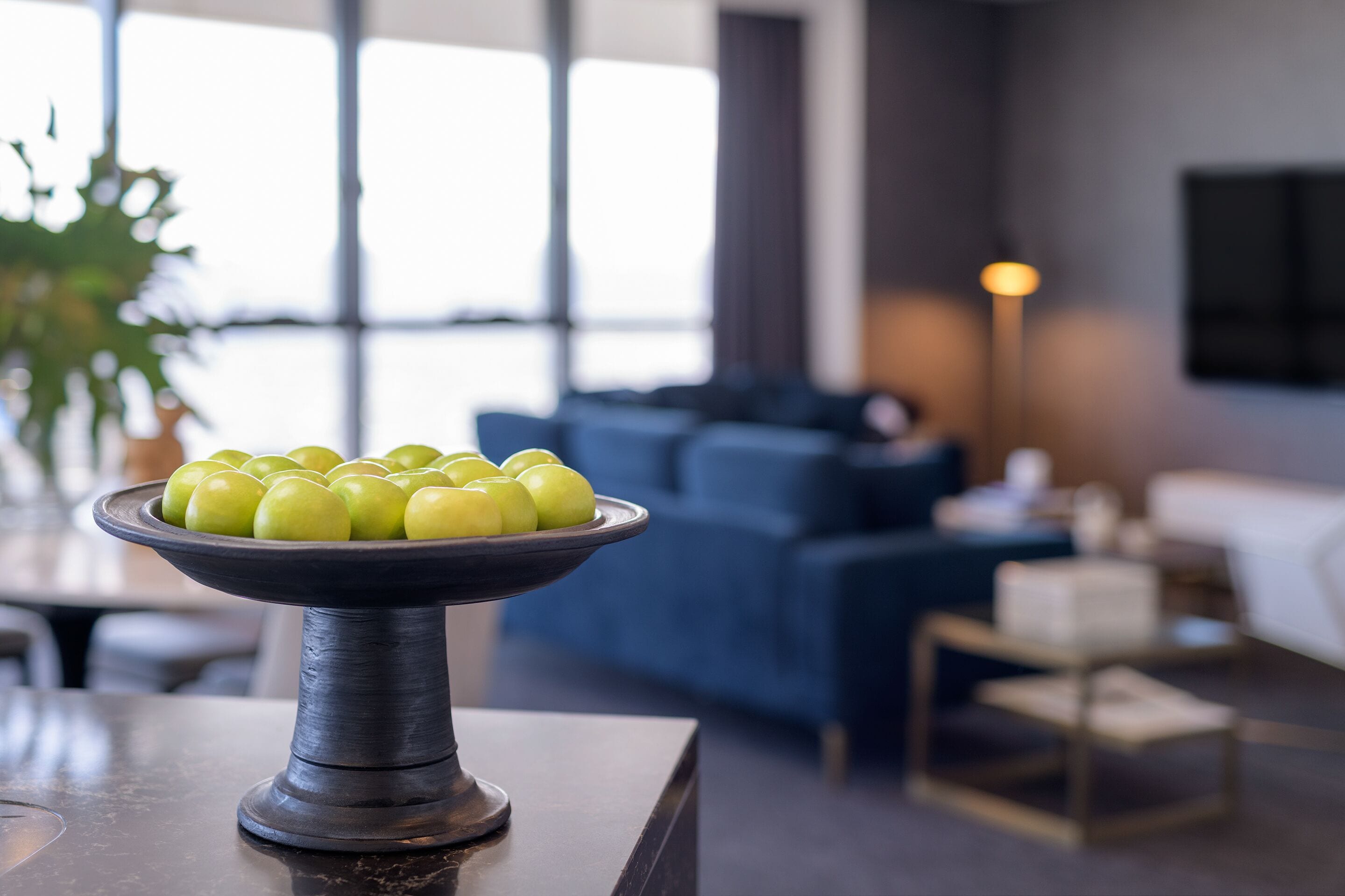 A modern living room with a dark-toned sofa, a coffee table with a bowl of green apples, and a large window providing natural light.