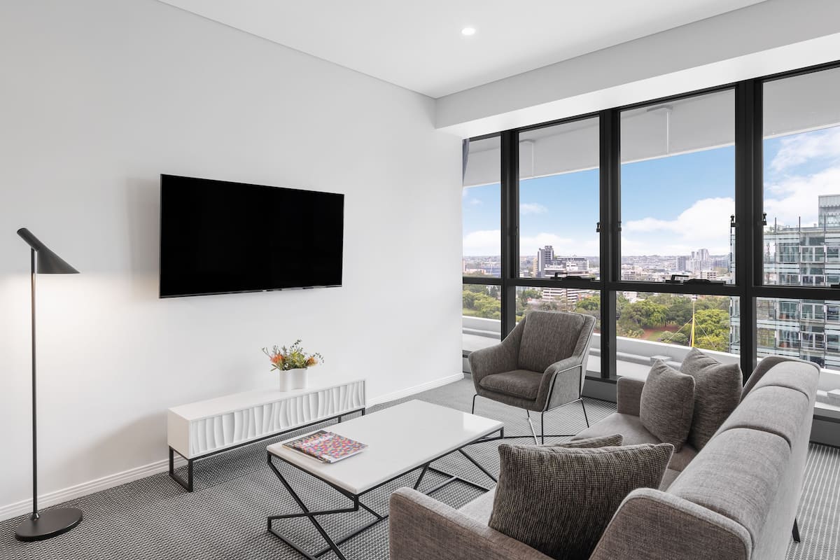 A modern living room with a large TV, comfortable seating, a coffee table, and a view of the city through large windows.