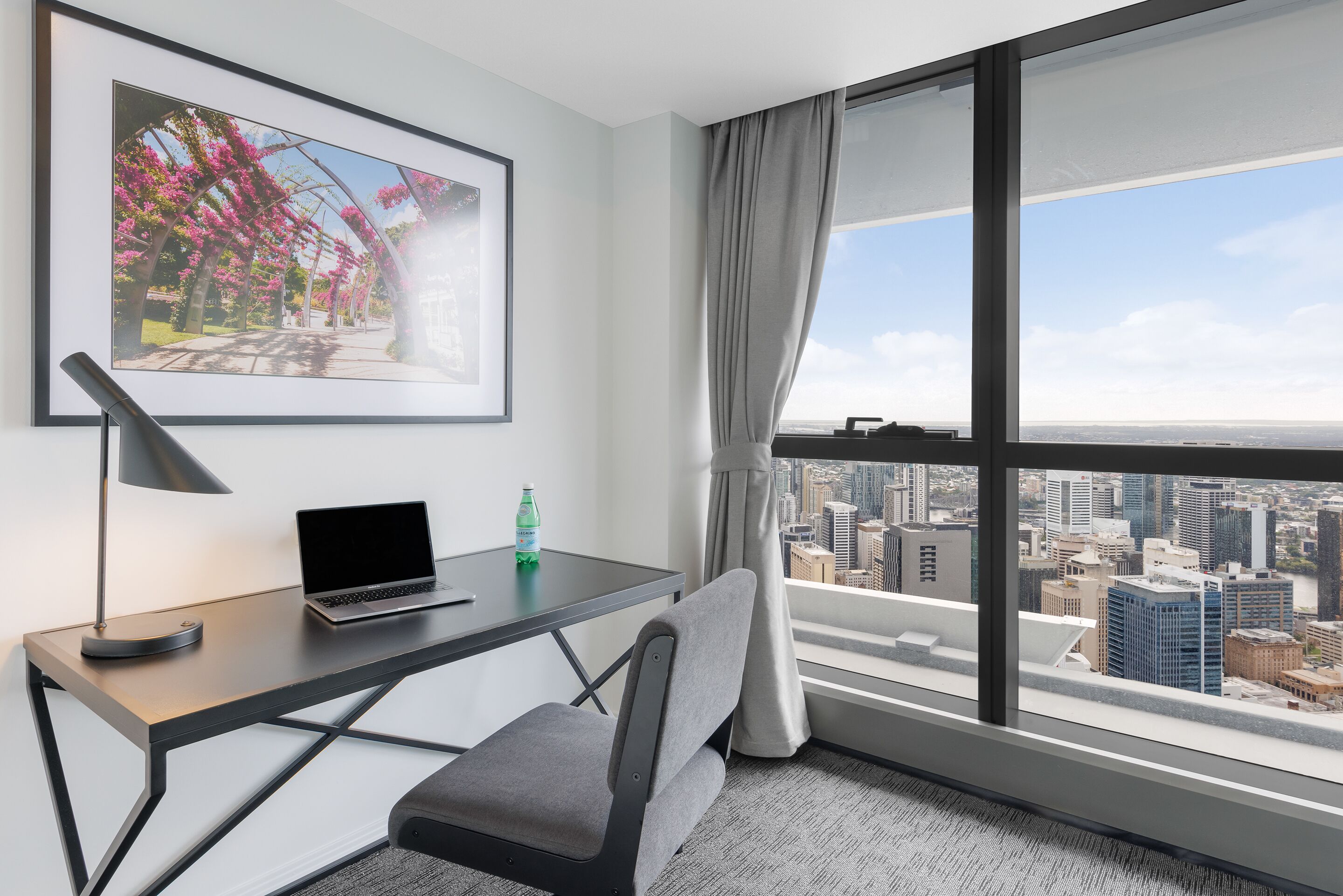 Habitación de hotel moderna con vista a la ciudad, un escritorio con una computadora portátil, una silla y un cuadro en la pared.