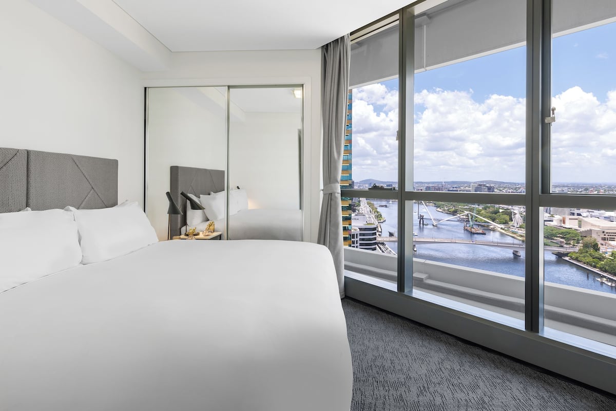 A hotel room with a large bed, a nightstand, a lamp, and a view of a cityscape through a large window.