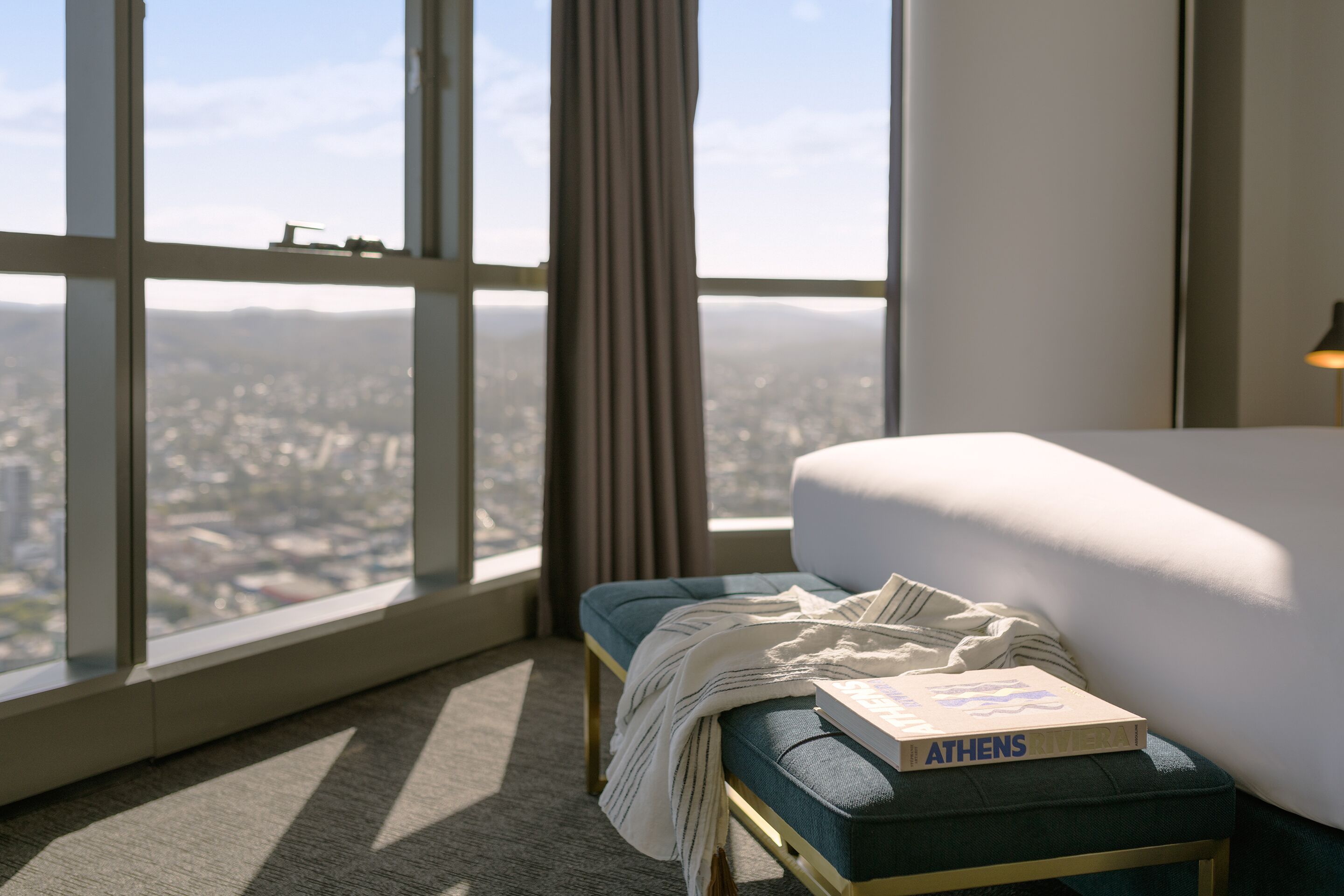Habitación de hotel con un ventanal amplio que ofrece vistas a la ciudad, una cama con edredón blanco, un banco tapizado en azul y un libro titulado 'ATHENS RIVIERA'.