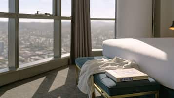 Habitación de hotel con un ventanal amplio que ofrece vistas a la ciudad, una cama con edredón blanco, un banco tapizado en azul y un libro titulado 'ATHENS RIVIERA'.
