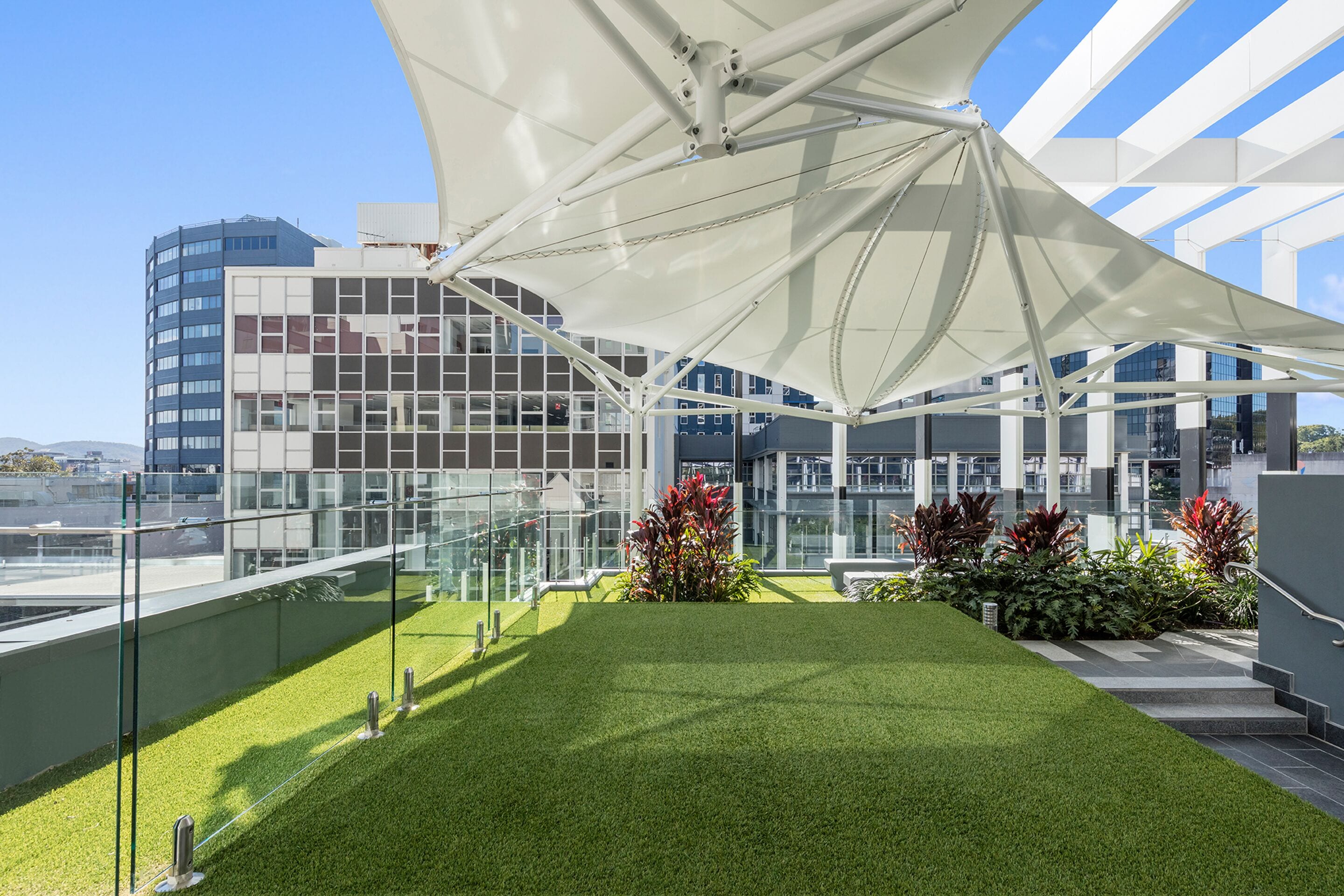 A rooftop terrace with artificial grass, a glass balustrade, and a modern building with a white canopy structure.