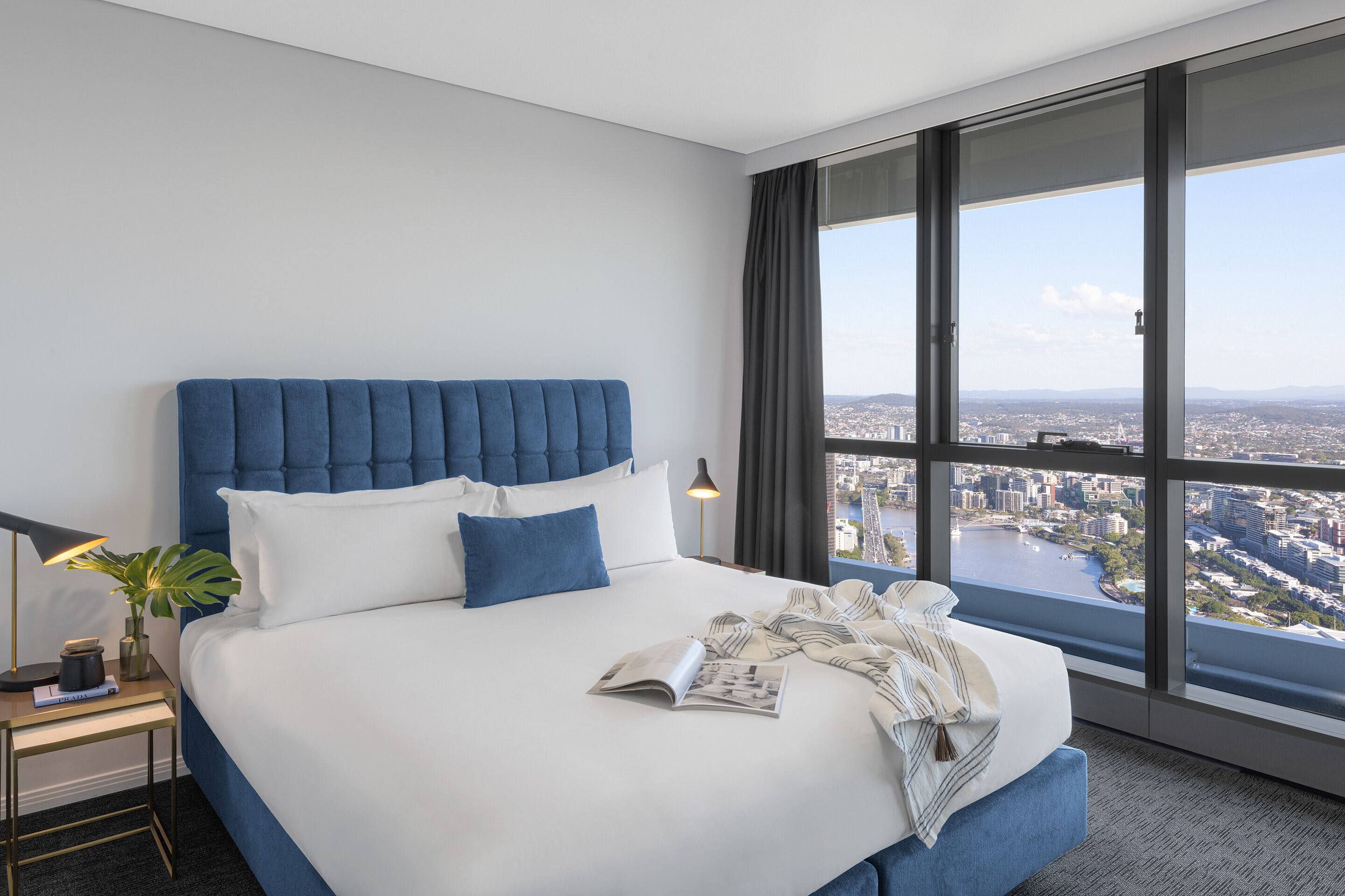 Habitación de hotel moderna con una cama grande, una mesita de noche, una lámpara y vistas a la ciudad a través de amplias ventanas.