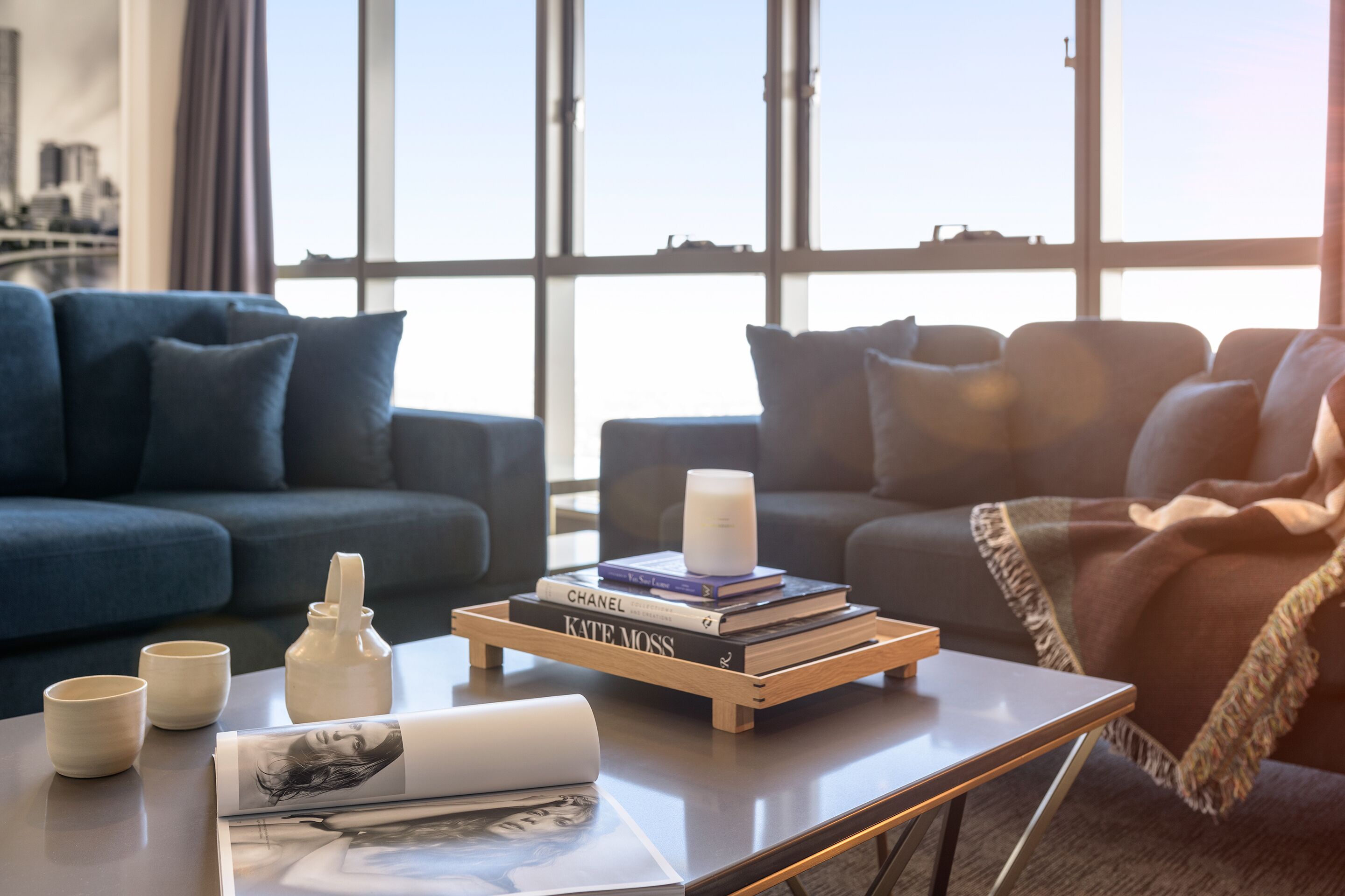 A modern living room with a sofa, a coffee table with books and a candle, and a view of the city through large windows.