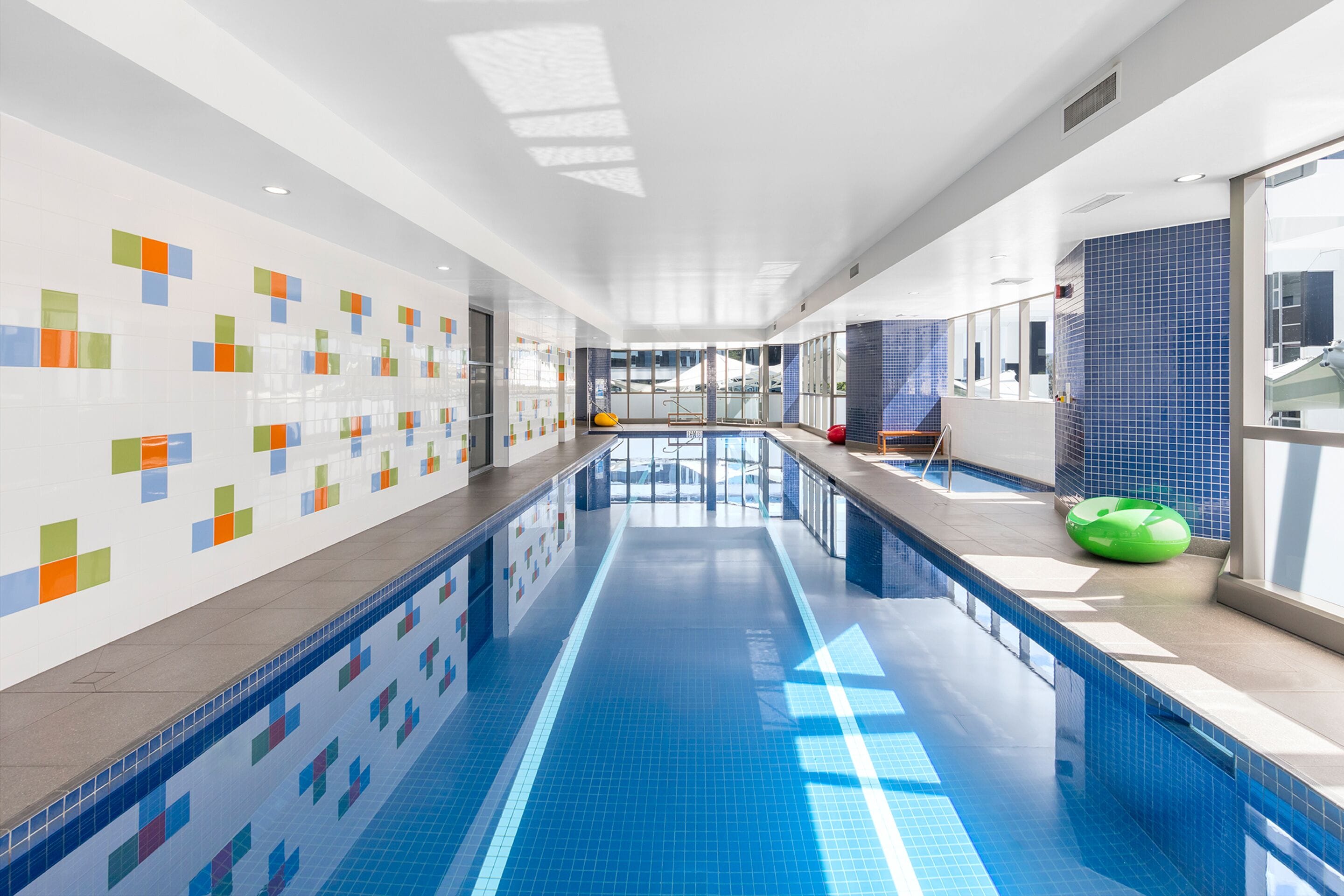 A modern indoor swimming pool with clear blue tiles, a long poolside area, and colorful wall tiles.