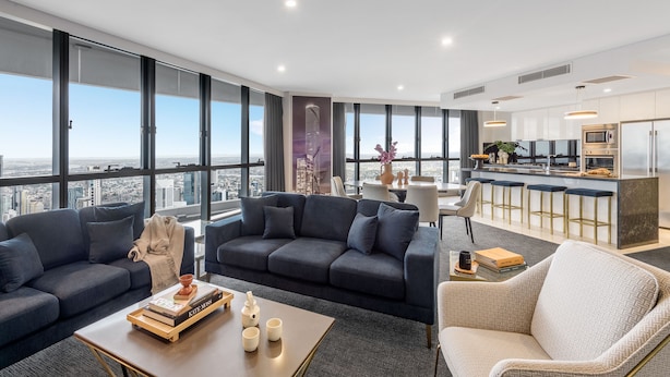Un salón moderno con un sofá gris oscuro, un sillón blanco y una mesa de centro con libros y un jarrón. Se observa una cocina con barra y ventanas amplias al fondo.