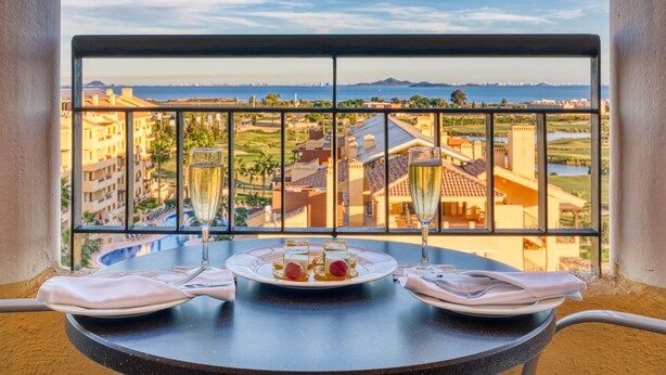 Una mesa de balcón con una copa de champán, un plato de comida y una servilleta. Se aprecia una vista que incluye edificios y una masa de agua.