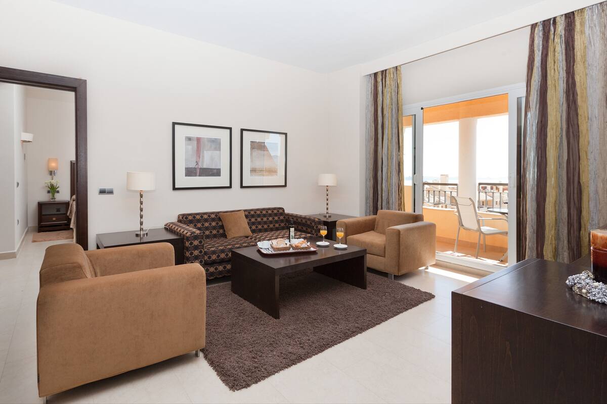 Een moderne woonkamer met een bank, fauteuils, een salontafel en een eethoek met uitzicht op een balkon.