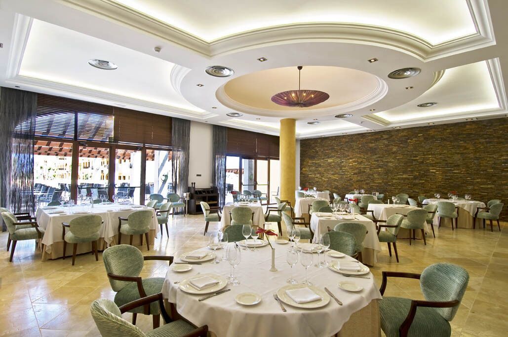 Un comedor amplio con mesas redondas dispuestas para varios comensales, con un column central y un fondo de pared de piedra.