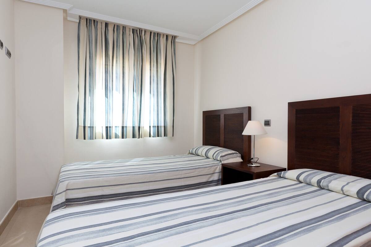 Een hotelkamer met twee bedden, een raam met gestreepte gordijnen, een bedlampje en een houten hoofdbord.