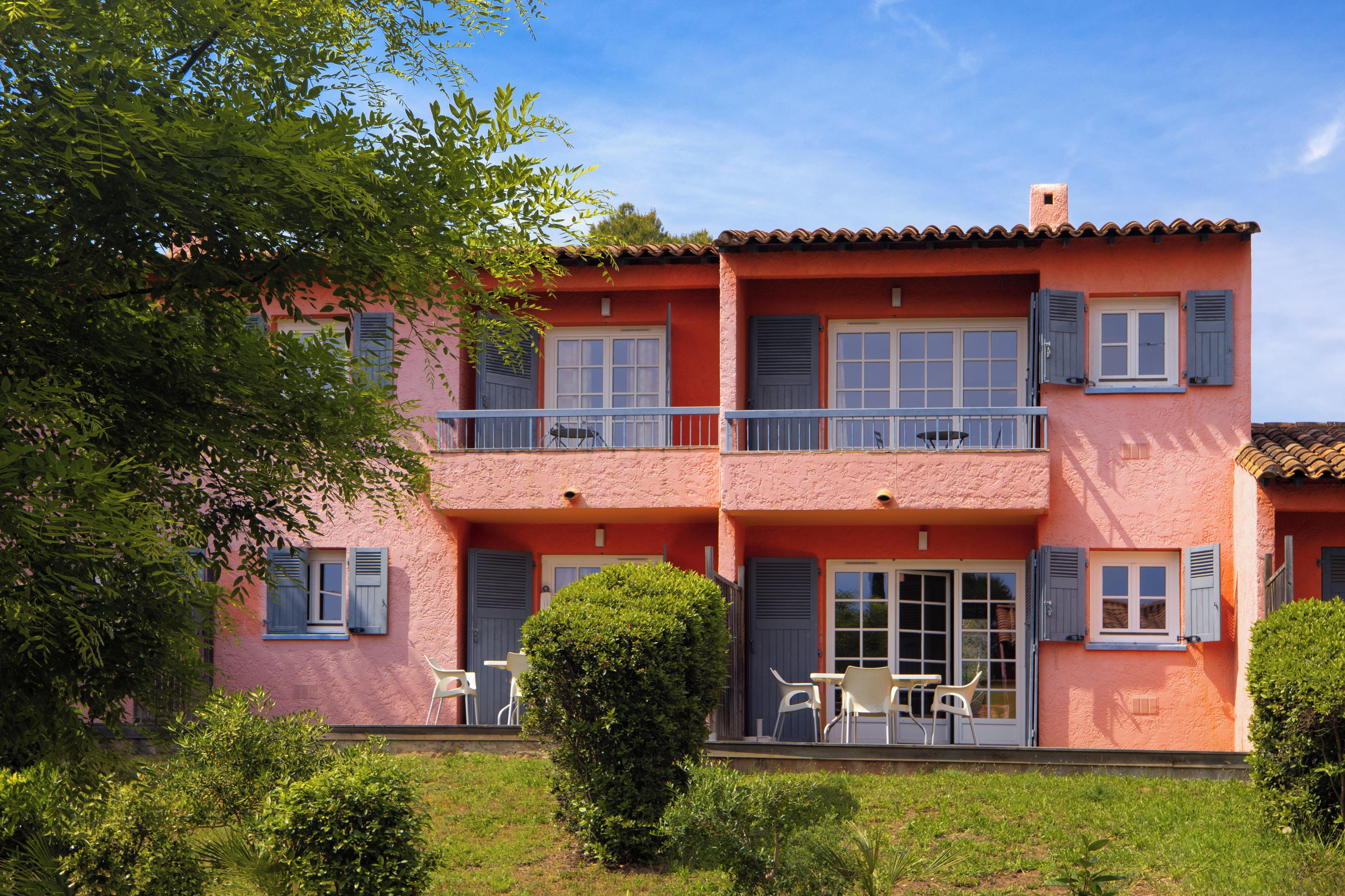 A two-story house with a balcony, a door with shutters, and a small garden area.