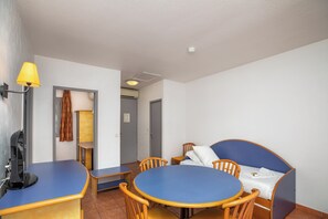 Una habitación con una mesa y sillas azules, una cama con cabecera azul, una lámpara y una puerta que da a otra habitación.