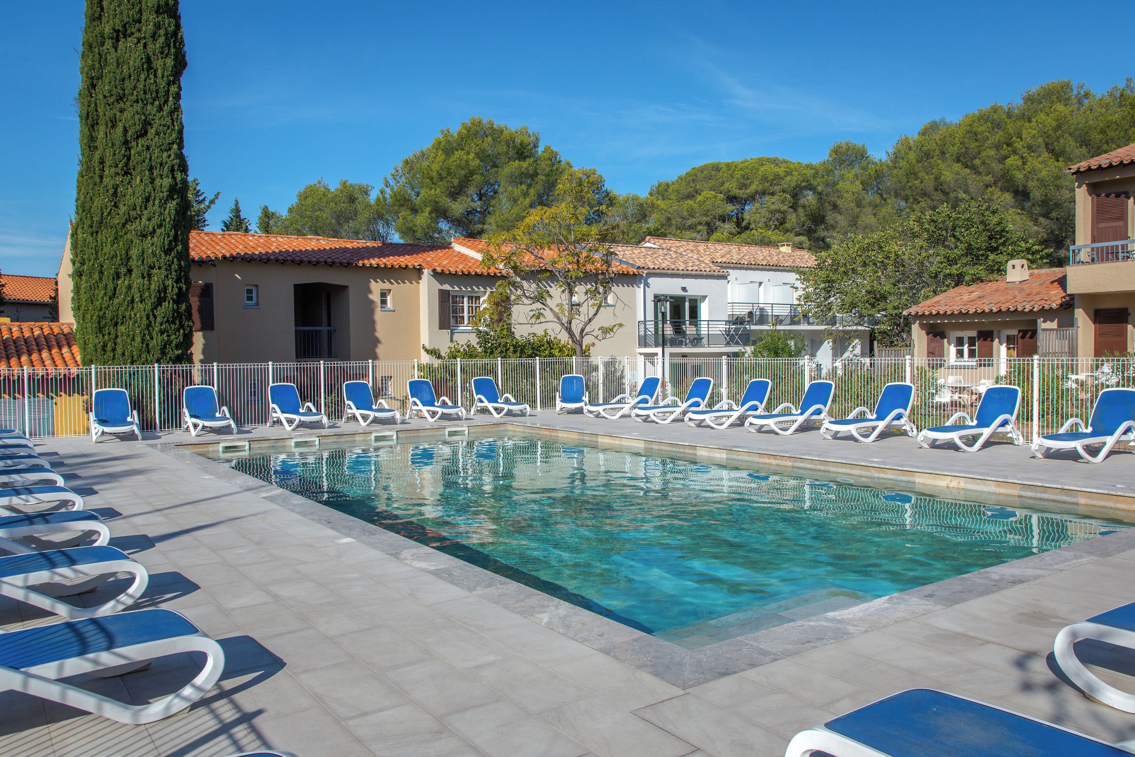 A swimming pool with lounge chairs and a fence, surrounded by buildings with tiled roofs.