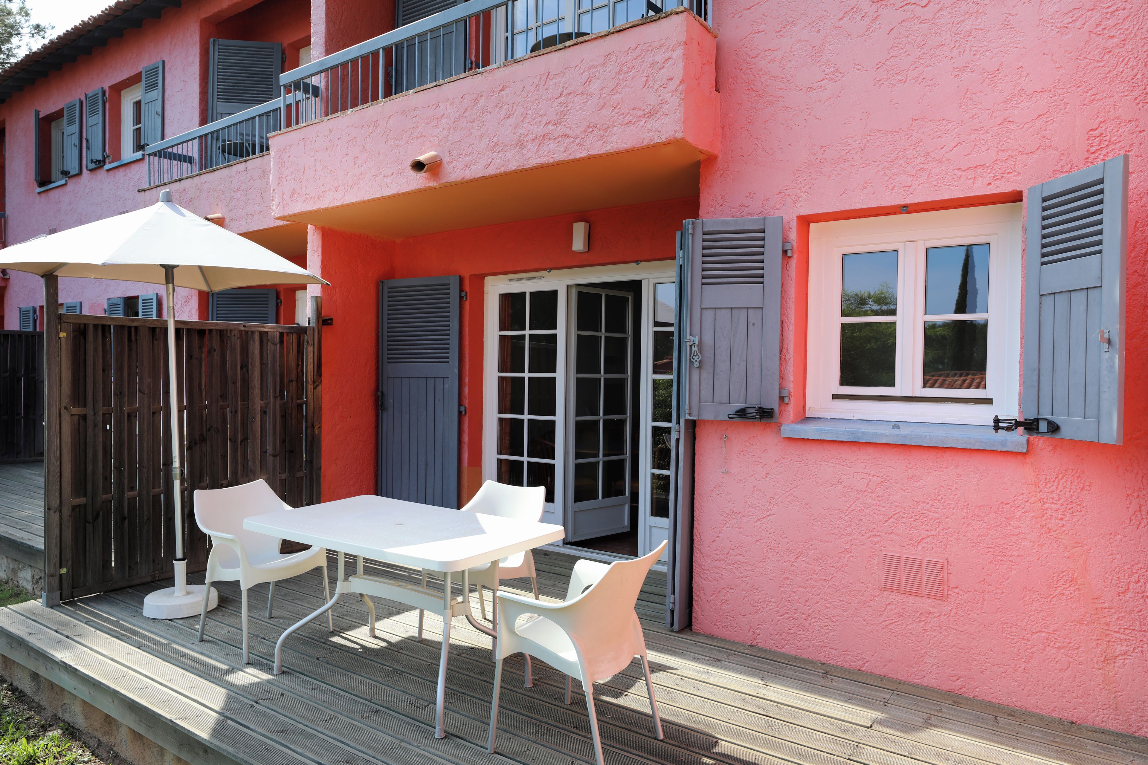 A pink building with a balcony, a white table with chairs, and a wooden deck.