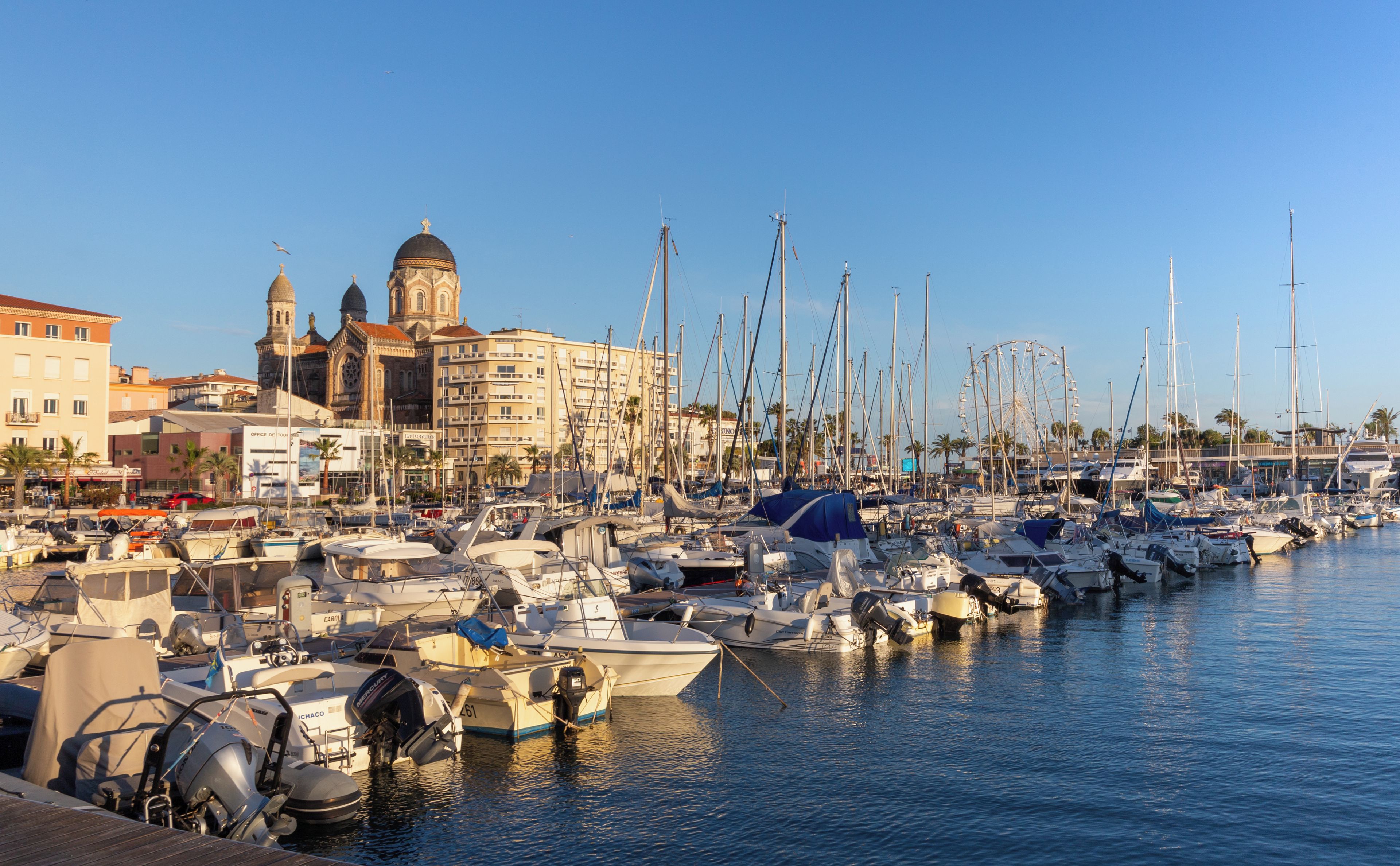 A marina with numerous boats docked, a prominent building with domed architecture, and a clear blue sky.