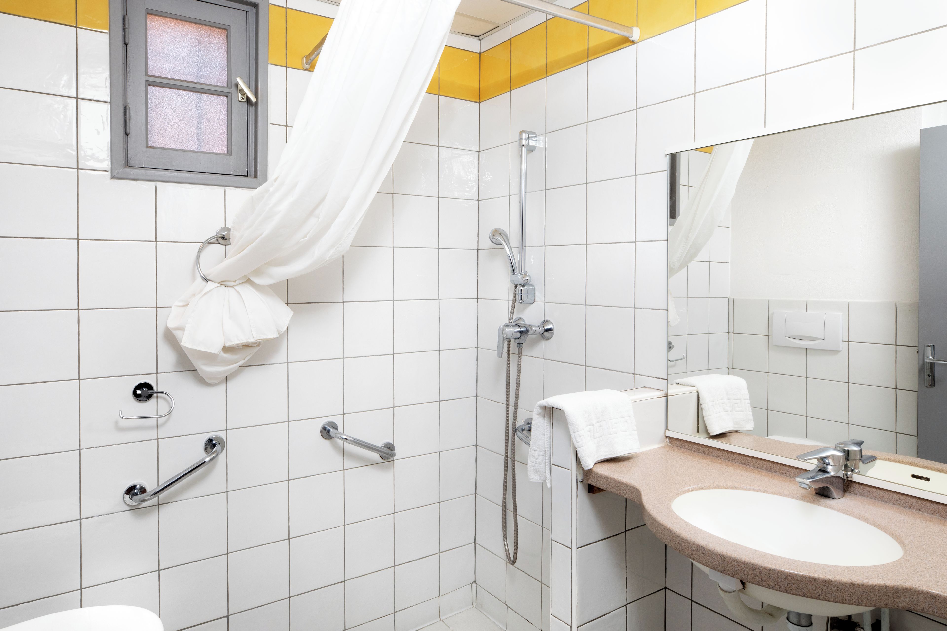 A bathroom with white tiled walls, a beige sink, a mirror, a shower with a handheld showerhead, and a curtain.