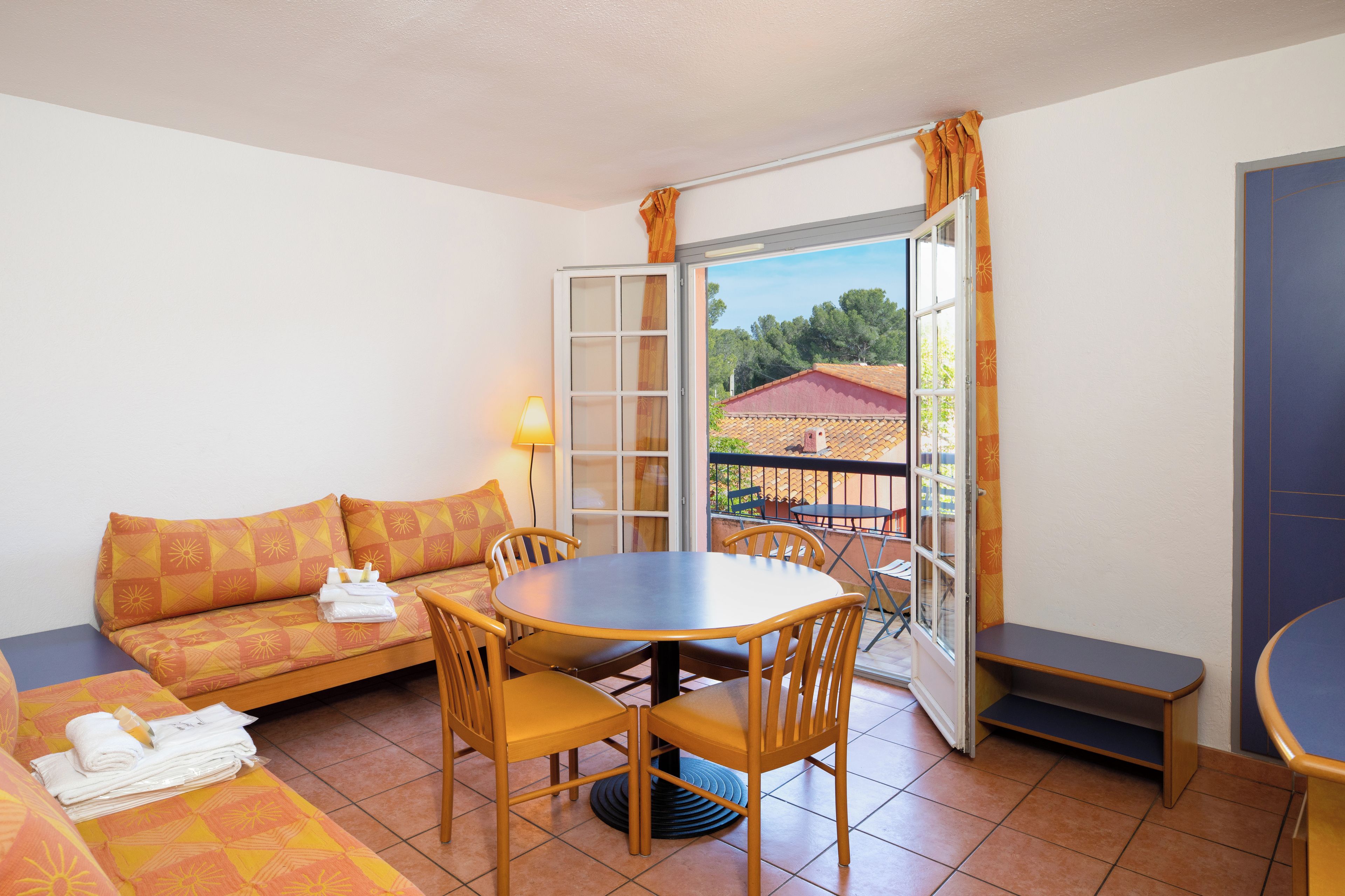 Una habitación con sofá, una mesa redonda y sillas. Hay una ventana con cortinas, y una lámpara sobre la mesa.