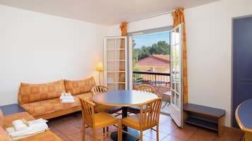 Una habitación con sofá, una mesa redonda y sillas. Hay una ventana con cortinas, y una lámpara sobre la mesa.