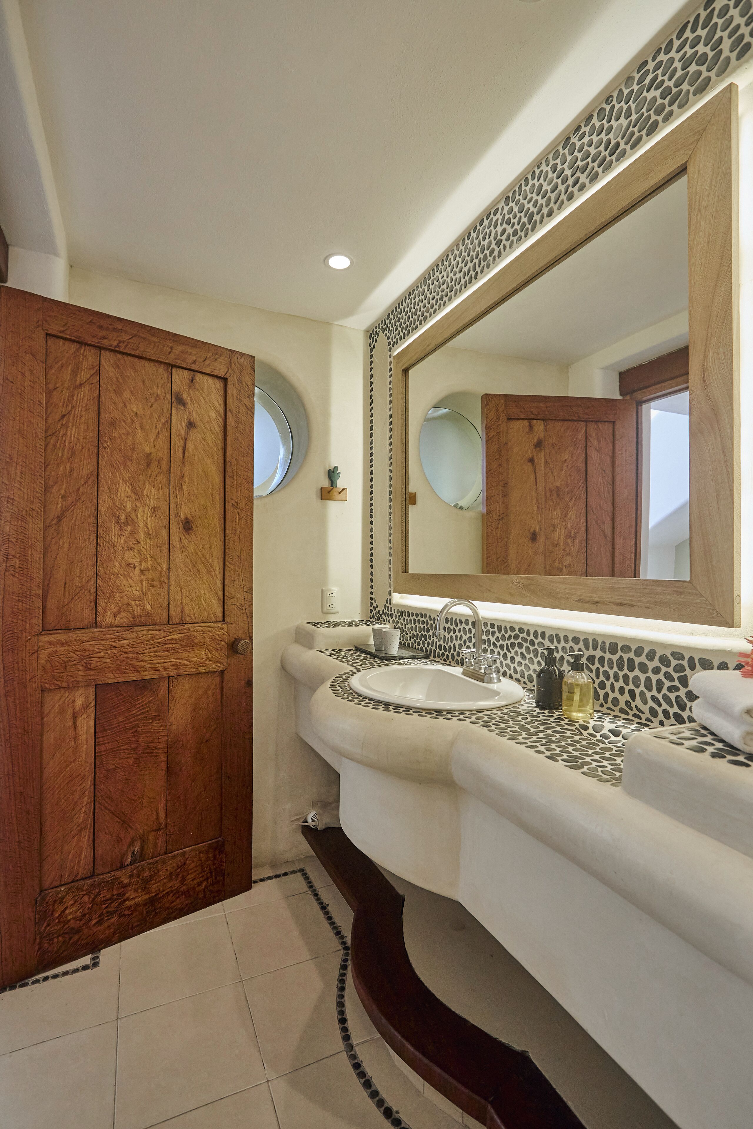 A bathroom with a wooden door, a round mirror, a white sink, and a tiled floor.
