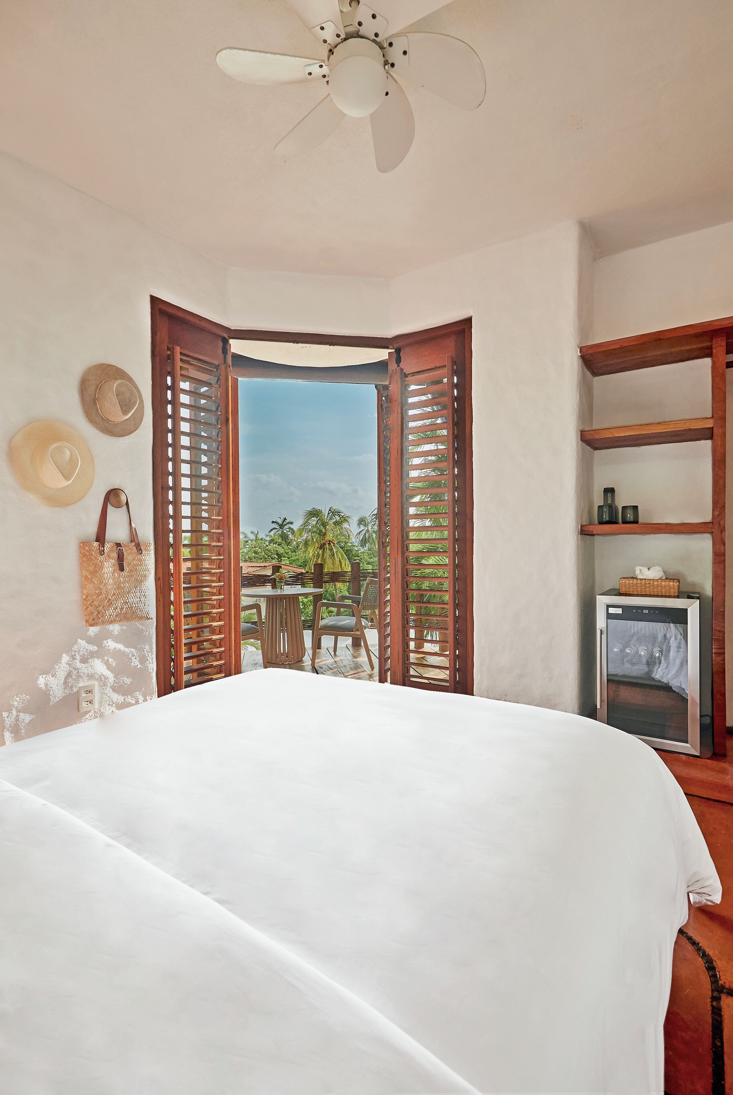 A bedroom with a bed, a ceiling fan, a wooden door leading to a balcony, and a small table with chairs.