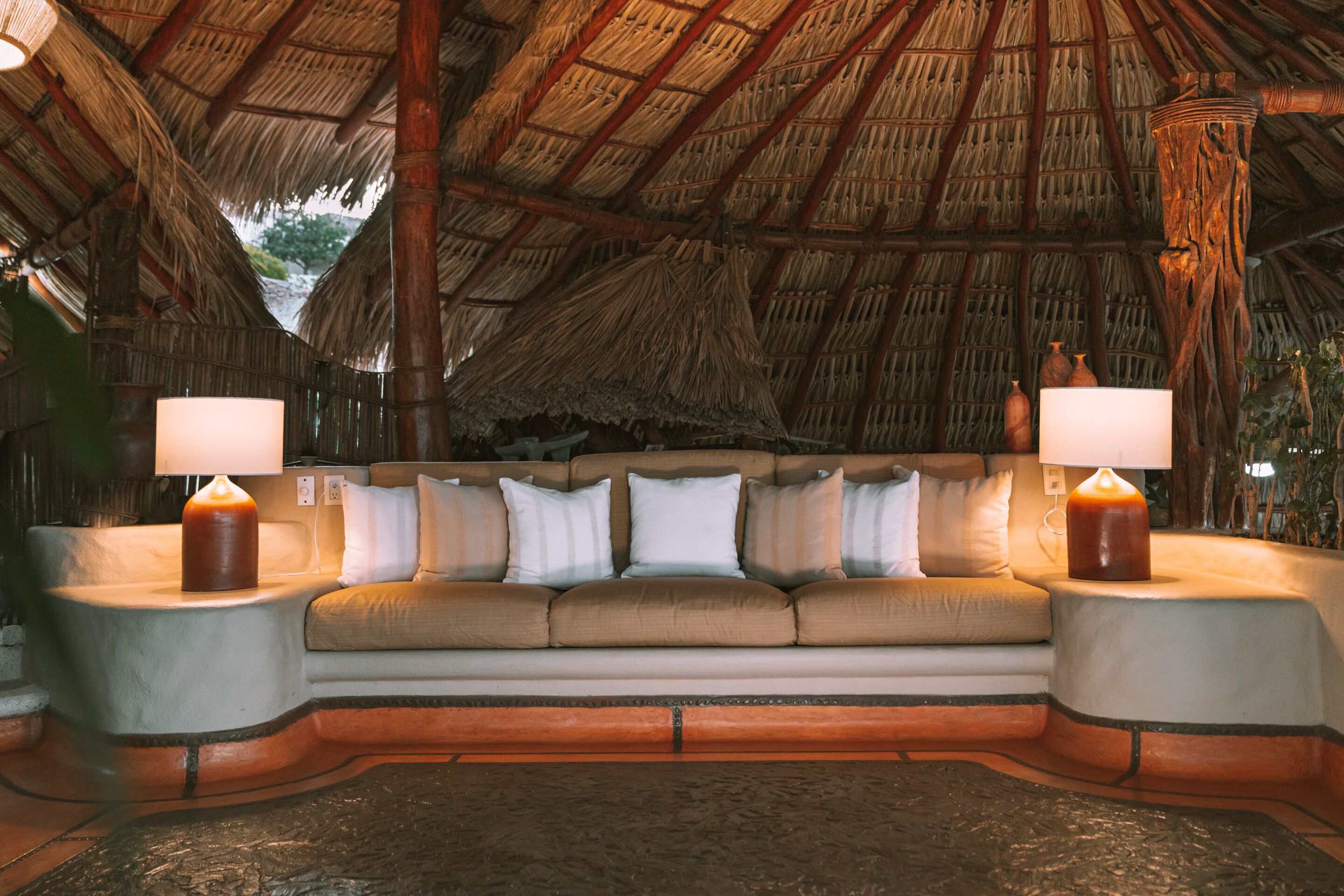 A room with a thatched roof, a beige sofa with white cushions, and two bedside lamps.