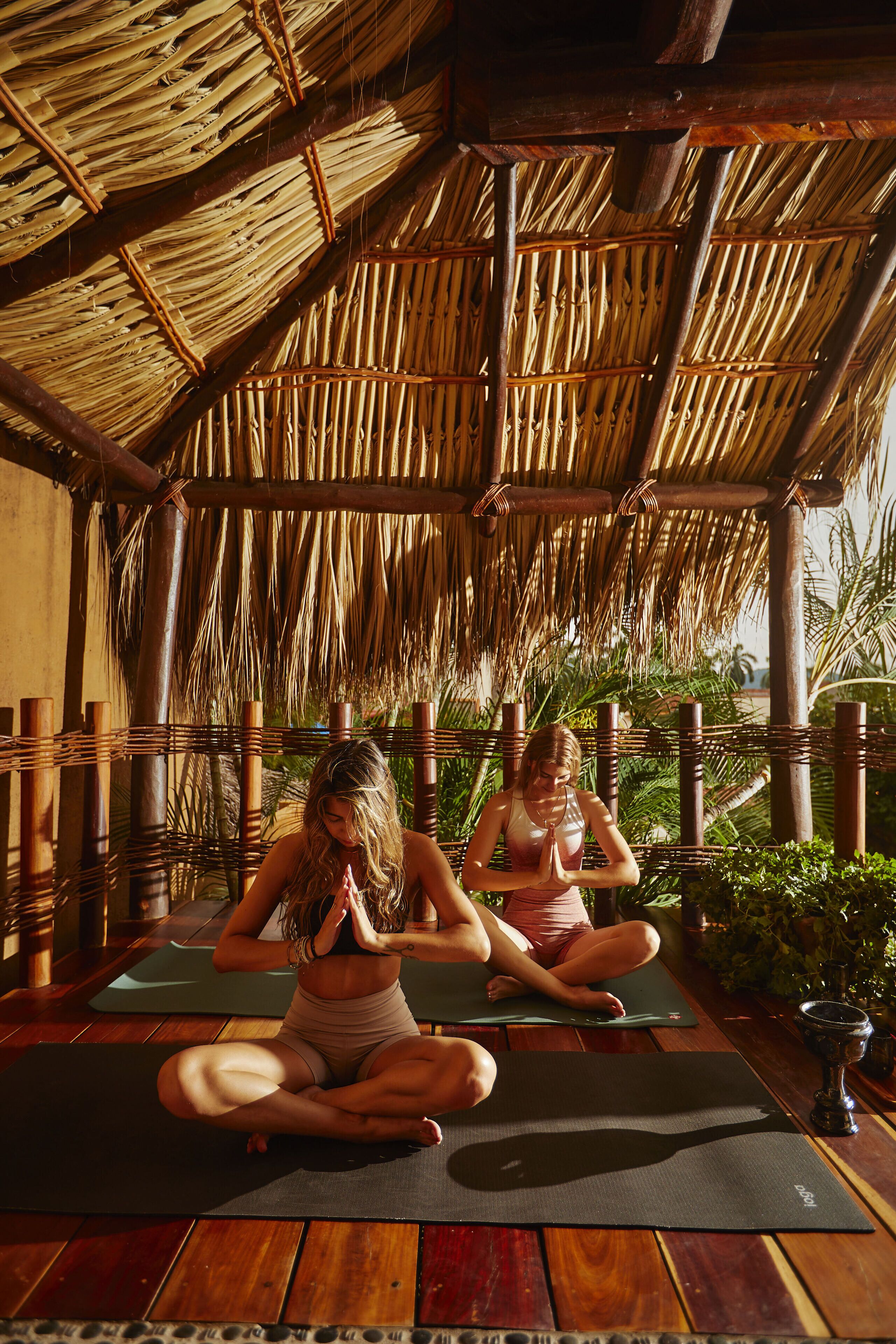 Dos personas sentadas sobre esterillas de yoga en una habitación con techo de paja y suelo de madera.