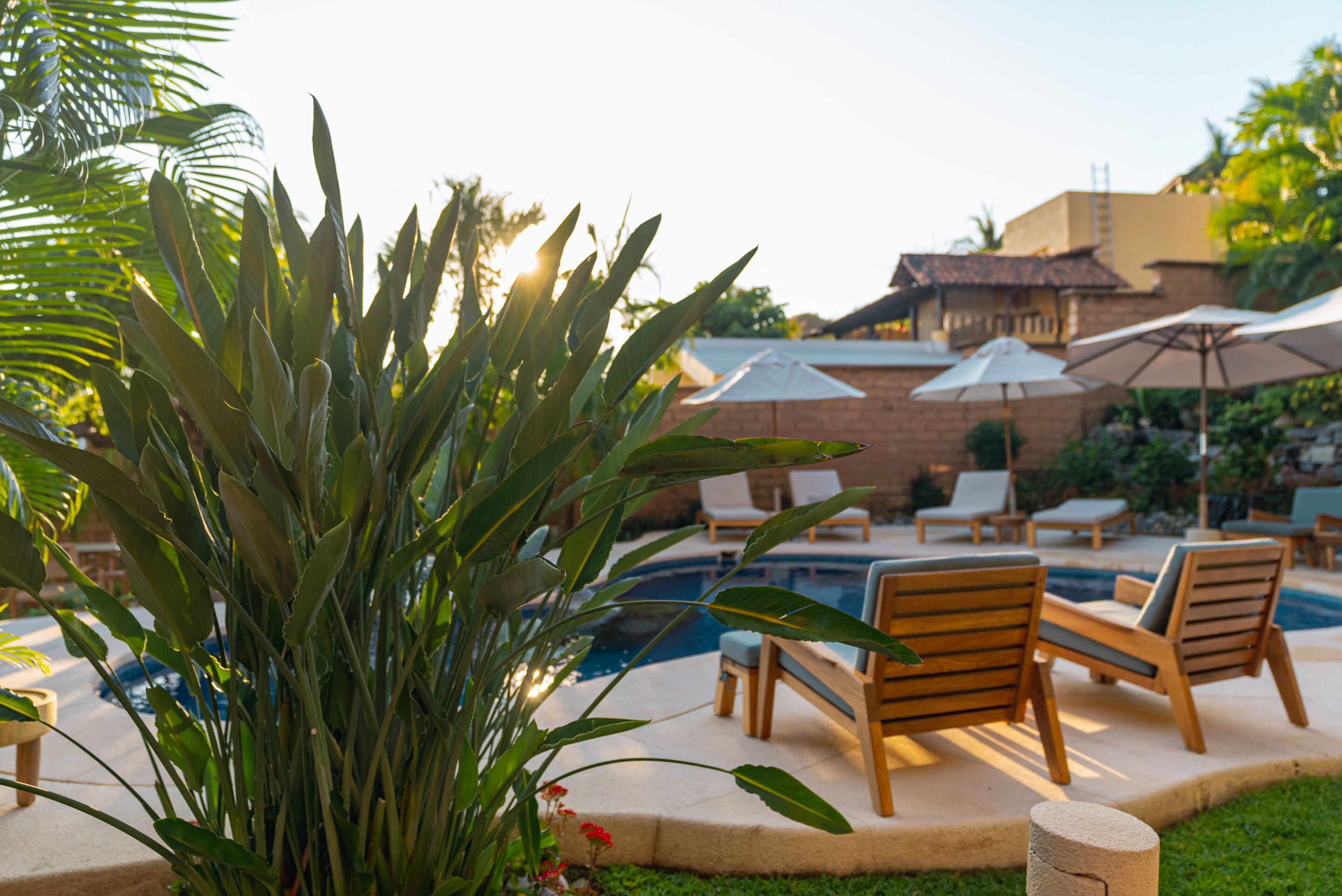 Zona junto a la piscina con sillones de descanso y sombrillas, rodeada de follaje y un edificio de ladrillo.