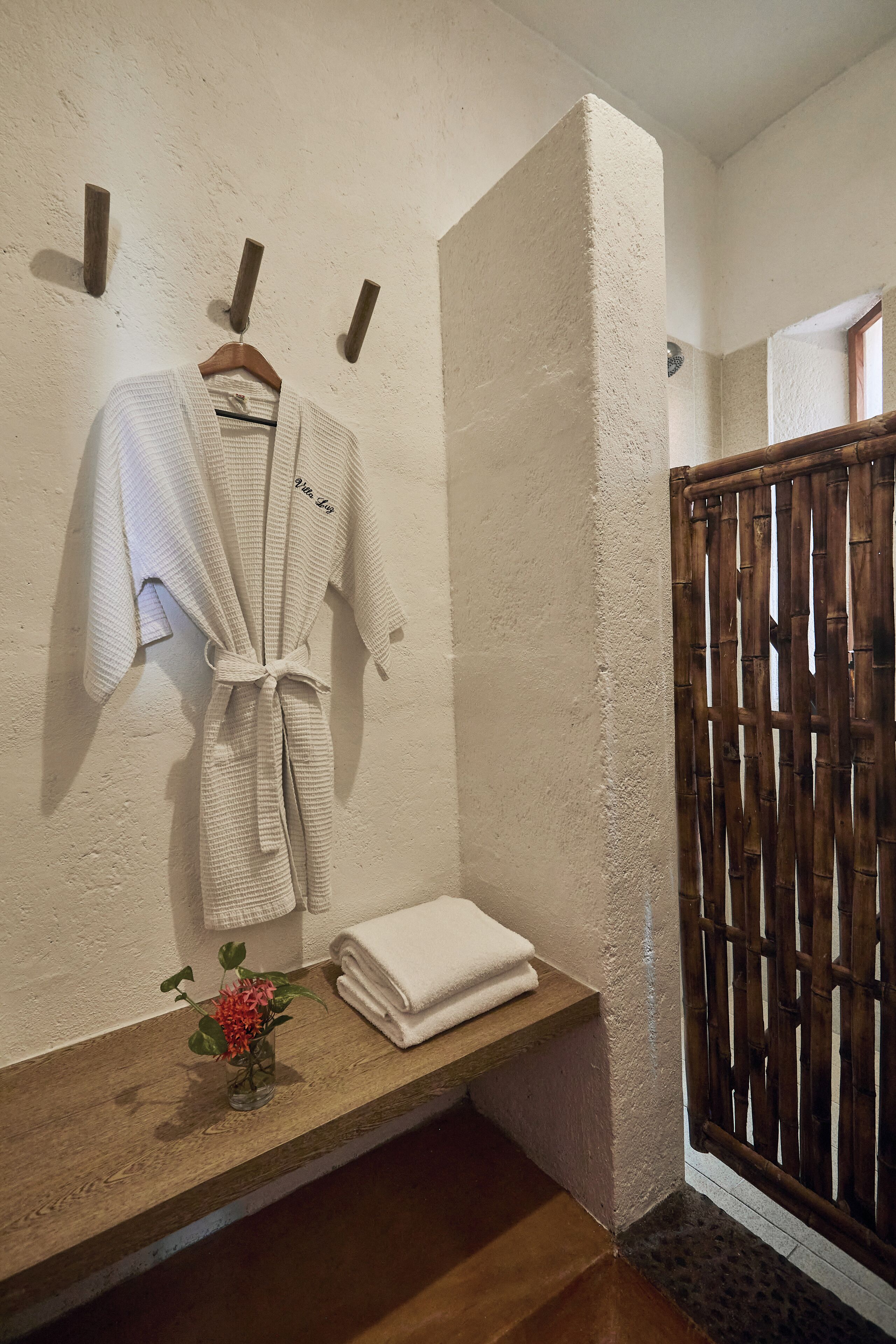Ein Badezimmer mit einer Holzbank, einem aufgehängten Bademantel und einer kleinen Vase mit Blumen.
