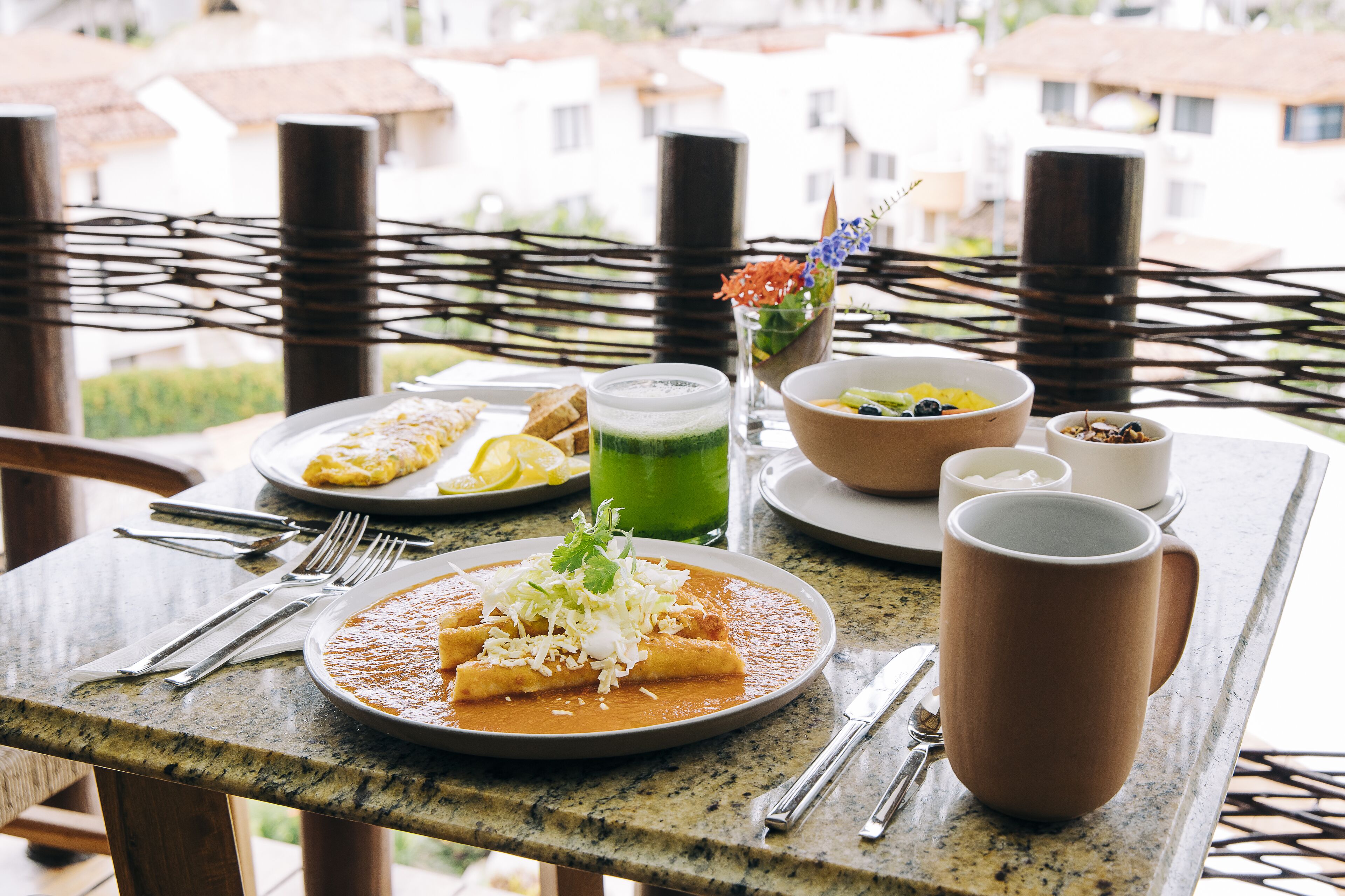 A table set with a plate of enchiladas, a bowl of fruit, a green drink, and a mug.