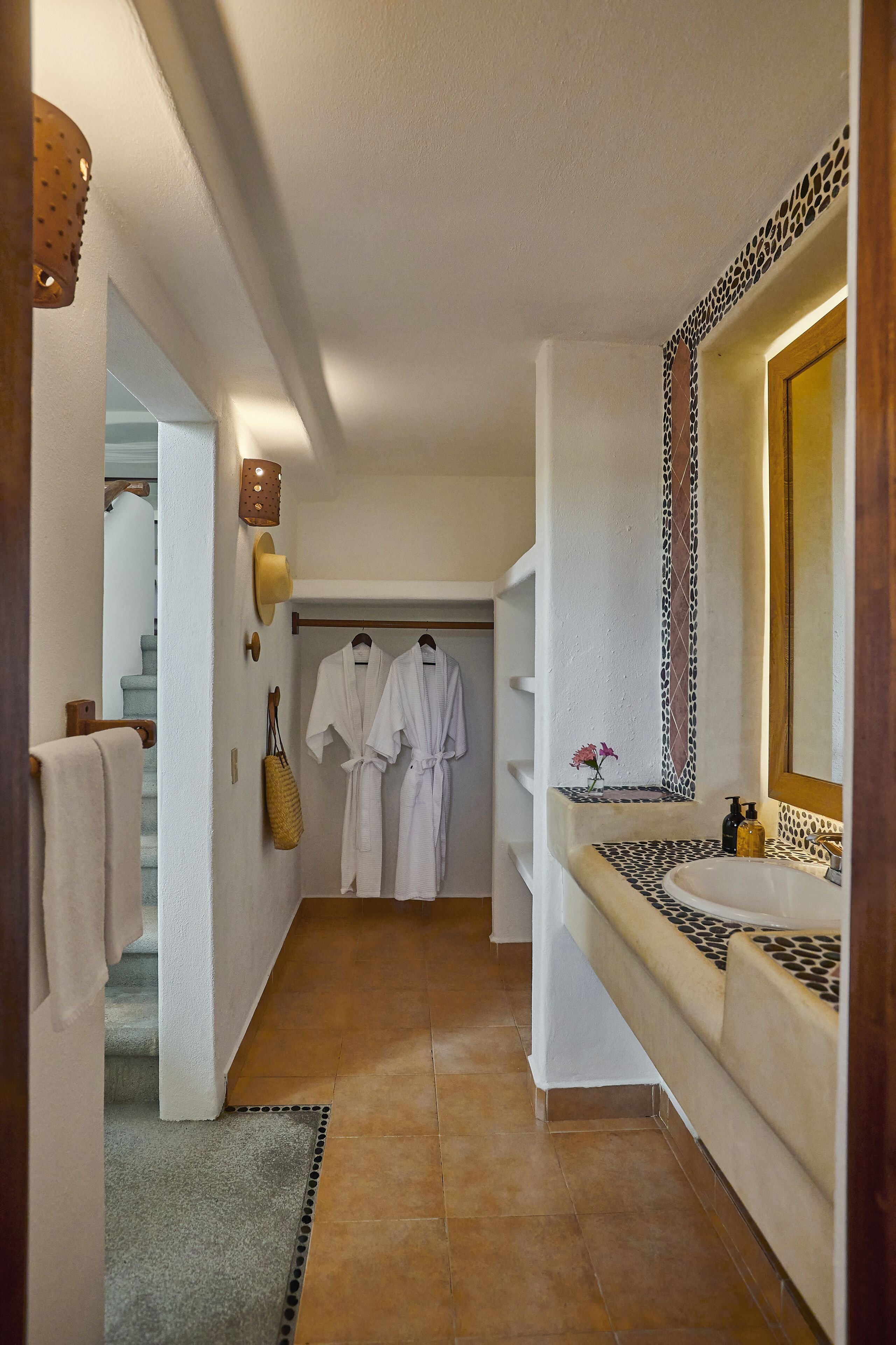 A hallway with a white wall, a tiled floor, a bench with a patterned seat, a mirror with a decorative frame, and a door leading to a room with a bed.