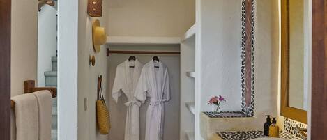 A hallway with a white wall, a tiled floor, a bench with a patterned seat, a mirror with a decorative frame, and a door leading to a room with a bed.