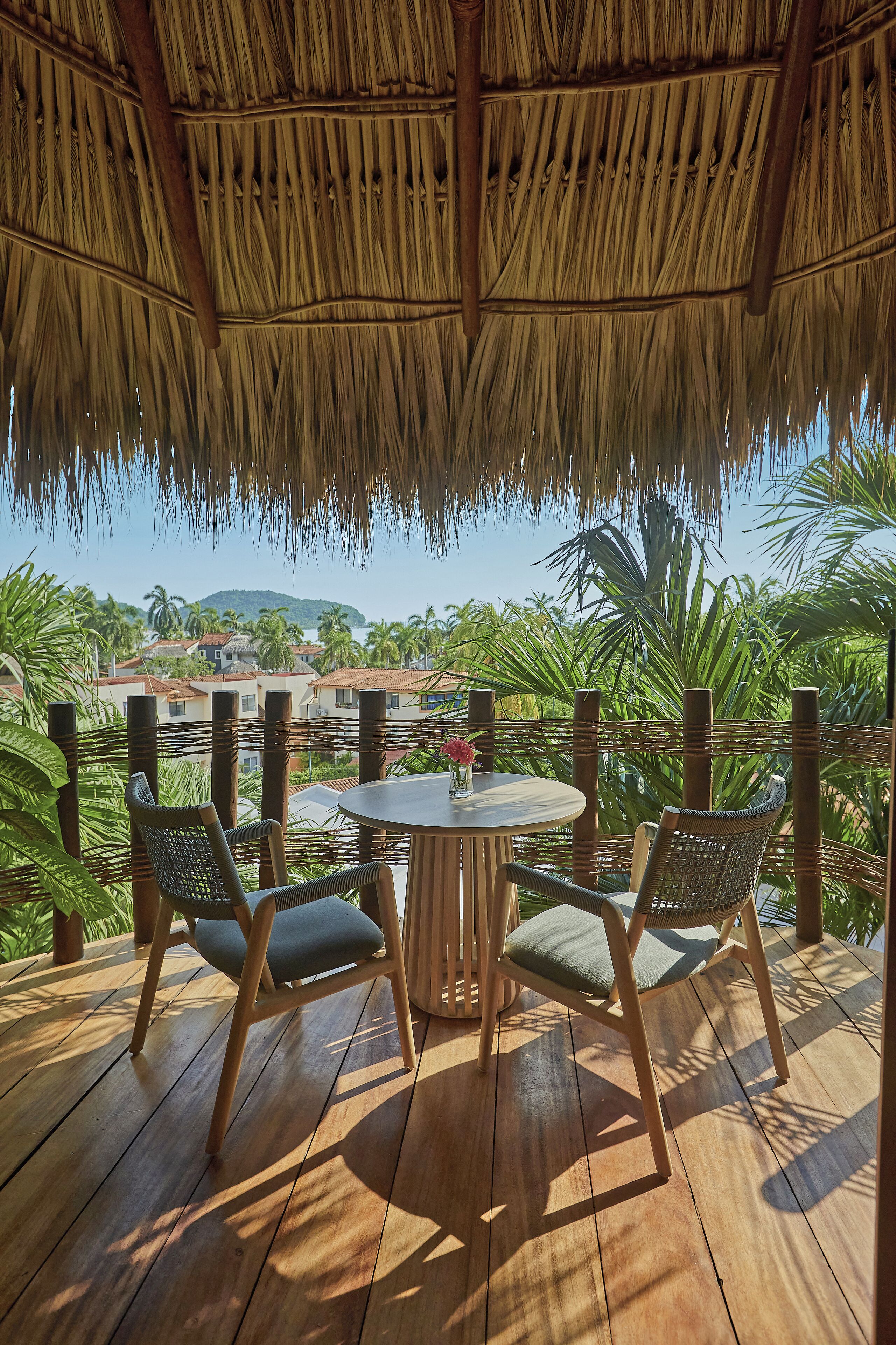 Une terrasse en bois surmontée d’un toit de chaume, avec une table ronde et deux chaises, donnant sur une zone côtière bordée de palmiers et de bâtiments.