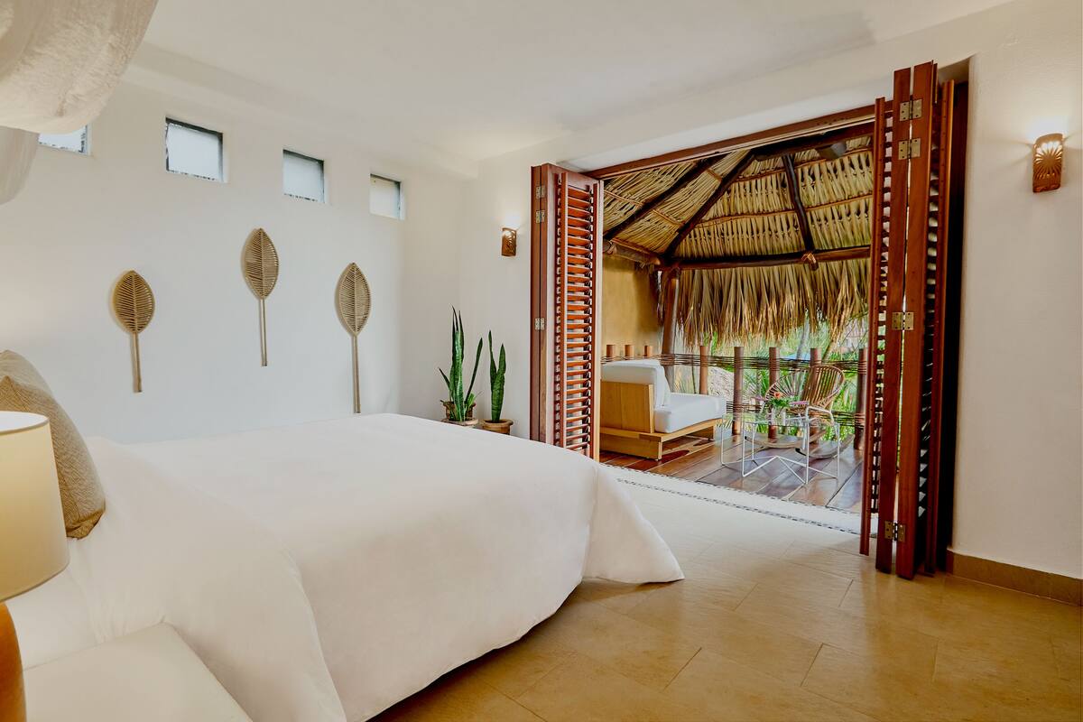 Een slaapkamer met een groot bed, een klein tafeltje met een lamp, een raam en uitzicht op een terras met stoelen en planten.