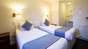 Ein Hotelzimmer mit zwei Betten, jeweils mit Kopfkissen und Decke. Es gibt einen Nachttisch mit Lampe, eine Kommode und eine Tür zu einem Badezimmer.