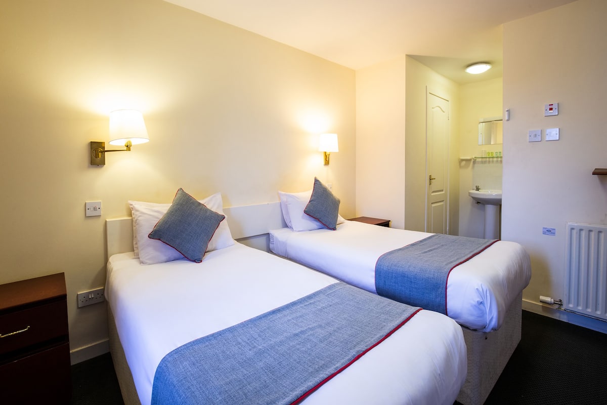 A hotel room with two beds, each with a pillow and a blanket. There is a nightstand with a lamp, a dresser, and a door leading to a bathroom.