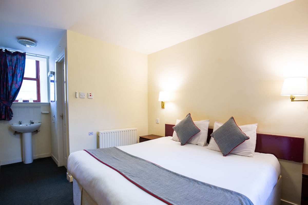 A hotel room with a large bed, a bedside table, a lamp, a window with curtains, and a bathroom with a sink and mirror.