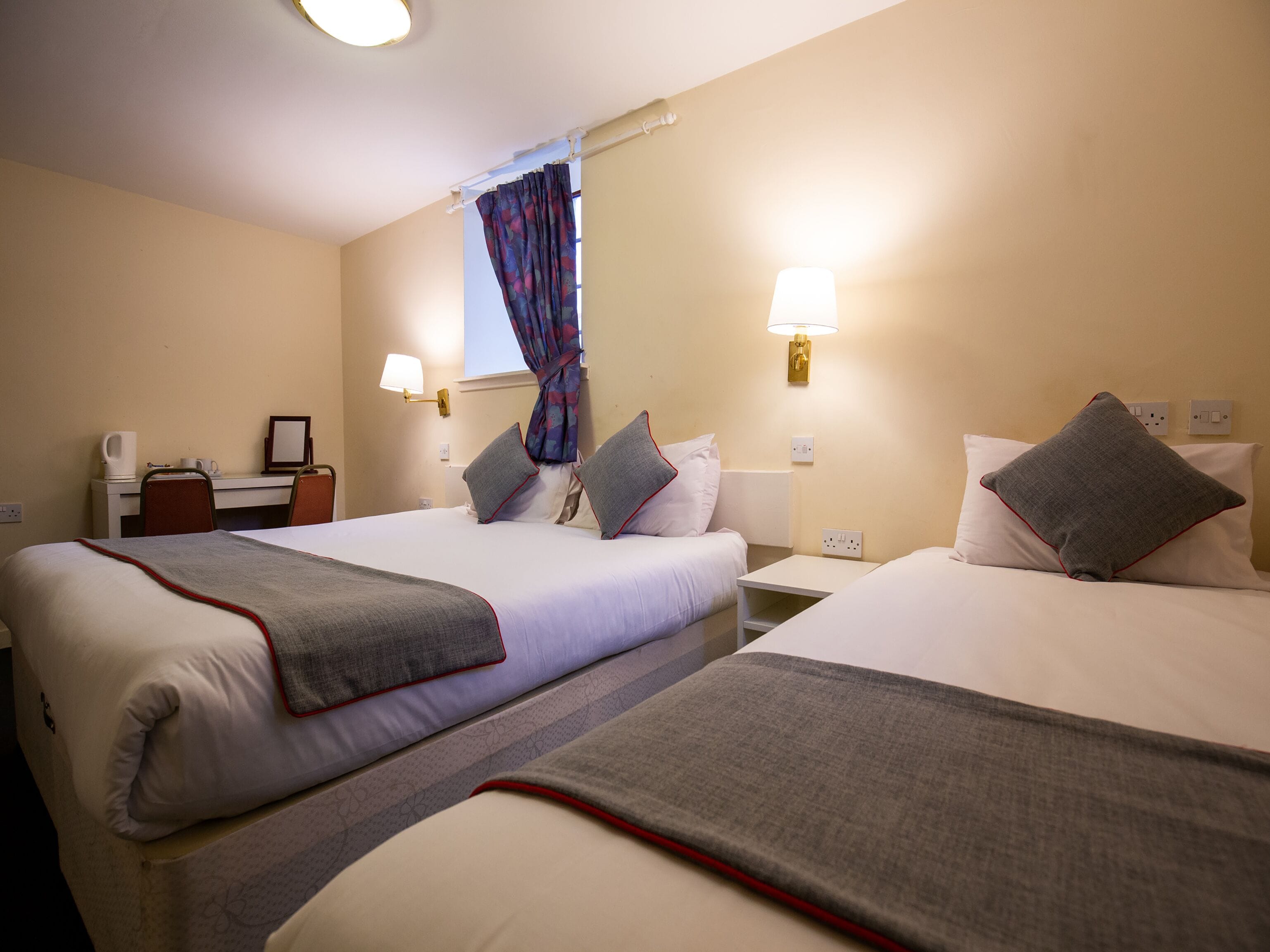 Une chambre d’hôtel avec deux lits, un bureau, une chaise, une fenêtre avec des rideaux et des lampes fixées au mur.
