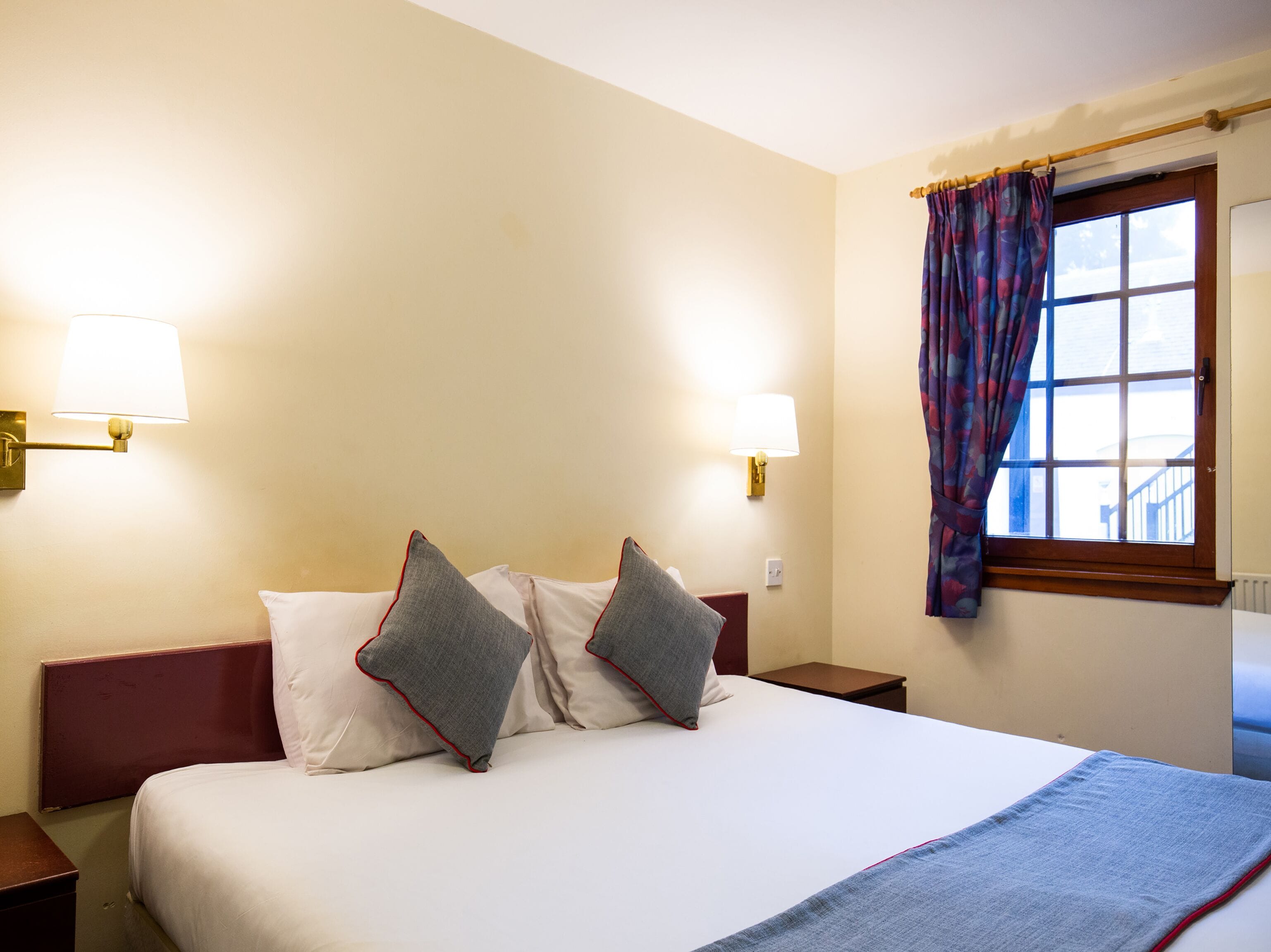 A hotel room with a large bed, two bedside lamps, a window with curtains, and a view of the outside.