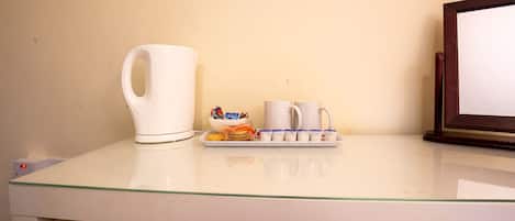 A white coffee maker, two white cups, a tray with snacks, and a framed picture on a glass table.