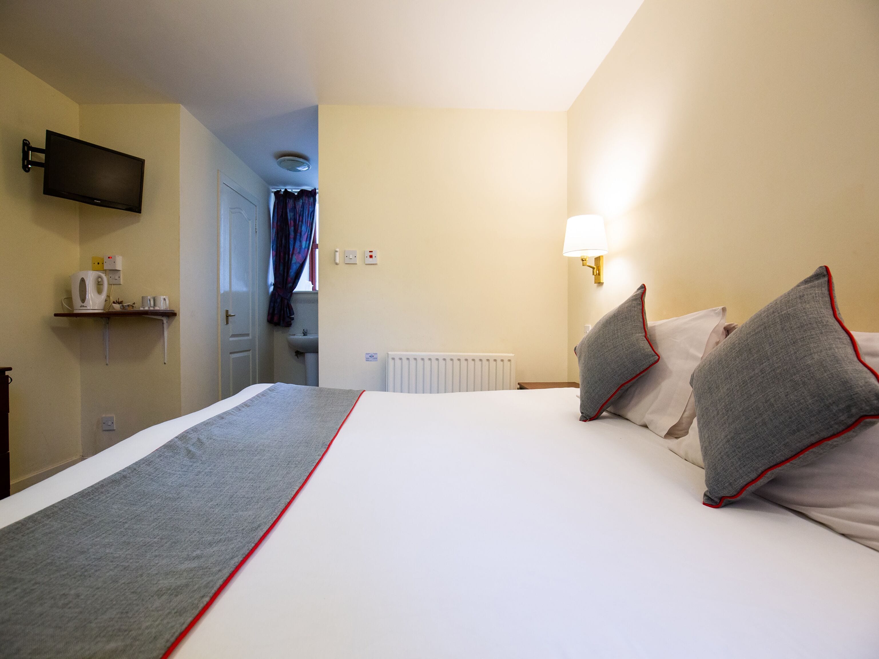 Habitación de hotel con una cama, un televisor montado en la pared, una lámpara de noche y un pequeño estante con una tetera y tazas.