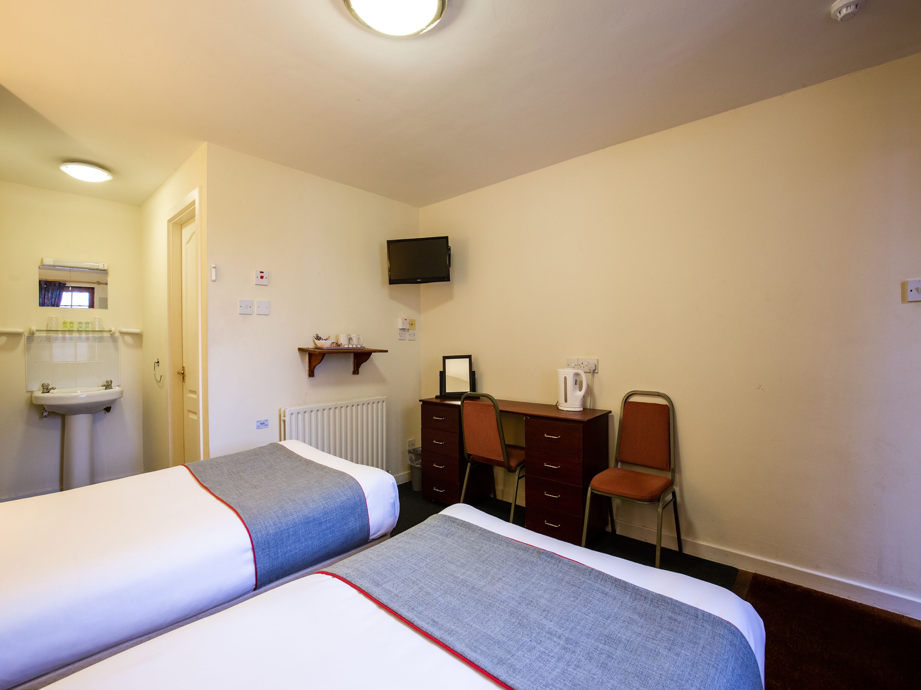 A hotel room with two beds, a desk, a chair, a television, a radiator, and a small shelf with toiletries.