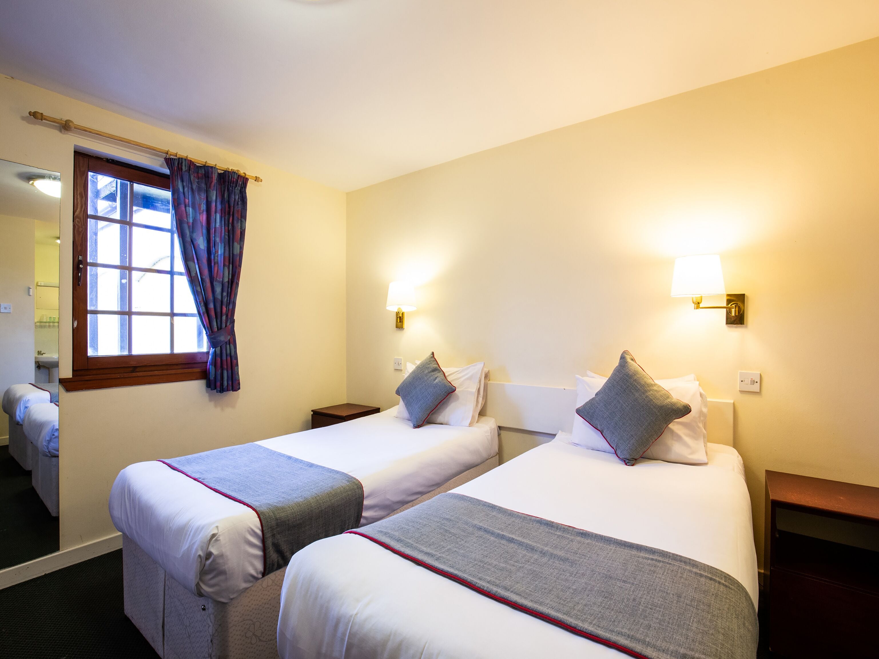 Habitación de hotel con dos camas, una ventana con cortinas, lámparas de noche y un baño visible al fondo.